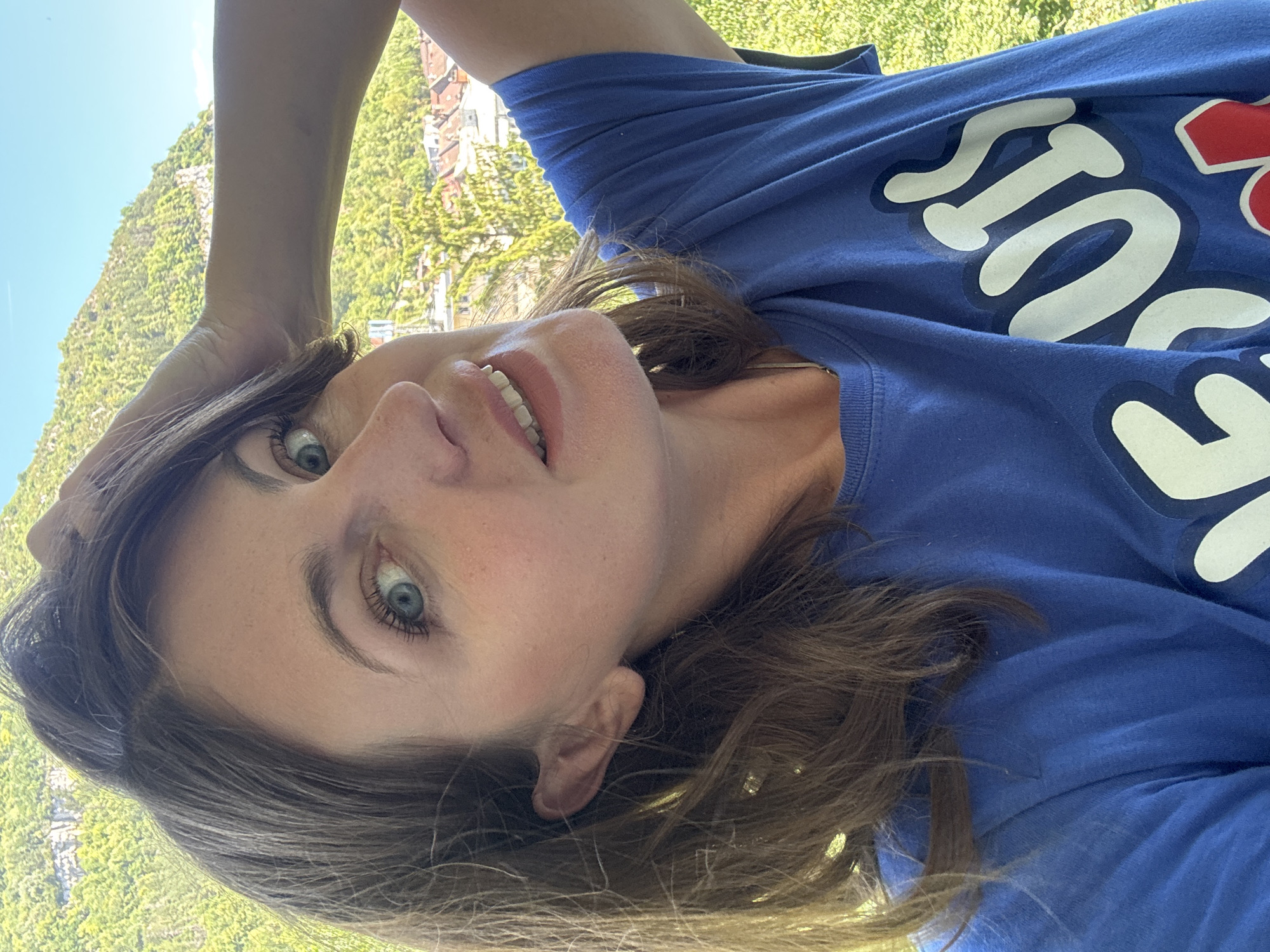 A woman is captured in a selfie, her face turned slightly to the side, with her right hand resting on top of her head, ruffling her brown hair. She is wearing a royal blue t-shirt with white and red lettering that reads "BOIS." Her blue eyes are looking towards the camera, and her mouth is open in a smile, revealing her teeth.
The background suggests an outdoor setting, possibly a balcony or elevated viewpoint, overlooking a town with red-tiled roofs nestled amongst green trees. Beyond the town, a steep, verdant mountain rises under a clear blue sky. This scene is consistent with the geography of Saint-Claude, France, a town situated in a valley with mountainous surroundings. The lighting indicates it is daytime, likely a sunny afternoon, casting soft shadows on the woman's face and highlighting the textures of the landscape.