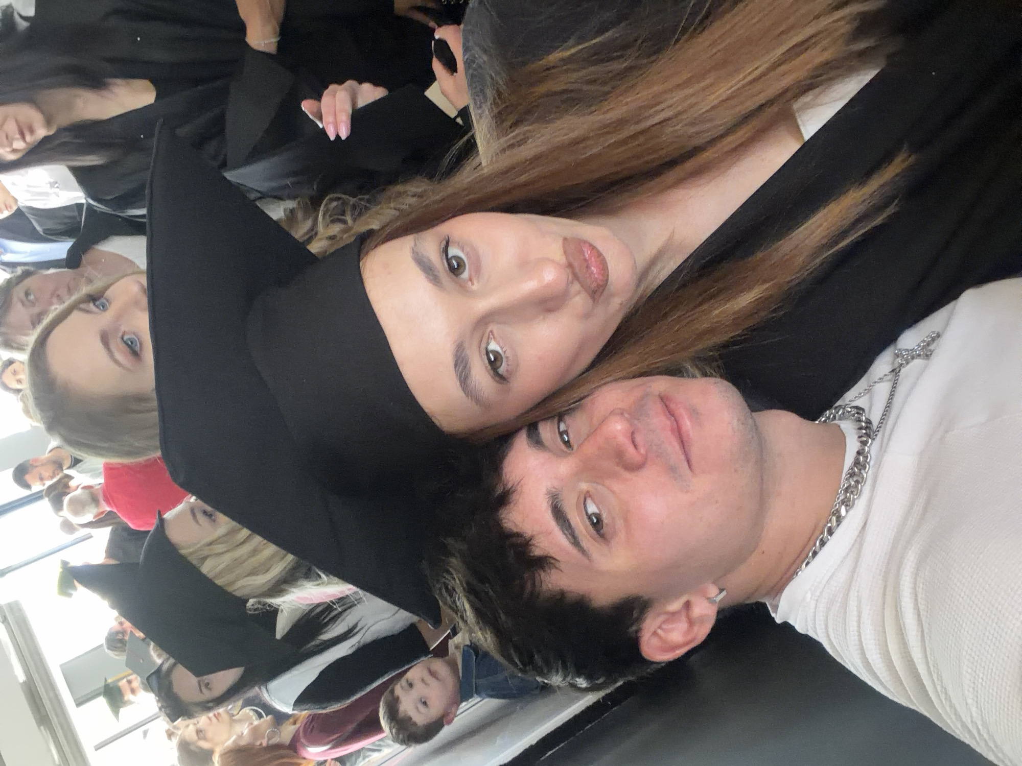 A selfie-style photograph captures two individuals in the foreground, with several others visible in the background, suggesting a gathering. The image is oriented with the subjects lying horizontally across the frame.
In the immediate foreground, a woman with long, light brown hair wears a black graduation cap and gown. She looks directly at the camera with a playful, pouty expression, her lips slightly pursed. Her eyes are light brown. Leaning against her is a man with short, dark hair. He is wearing a white, textured long-sleeved shirt and a thick silver chain around his neck. He has an earring in his left ear and a subtle smile, also looking towards the camera.
The setting appears to be an indoor space, likely a hall or event venue, brightly lit by natural light coming from windows or large openings in the background. Many other people are visible behind the main subjects, largely out of focus. Several of these individuals are also dressed in black graduation caps and gowns, indicating a graduation ceremony or a related celebration. One person with blonde hair is identifiable behind the main woman, and another wears a green graduation cap further back. A young boy is also partially visible in the lower right background.
The overall atmosphere is celebratory and joyful, consistent with a graduation event taking place in Plovdiv, Bulgaria. There is no visible text in the image.