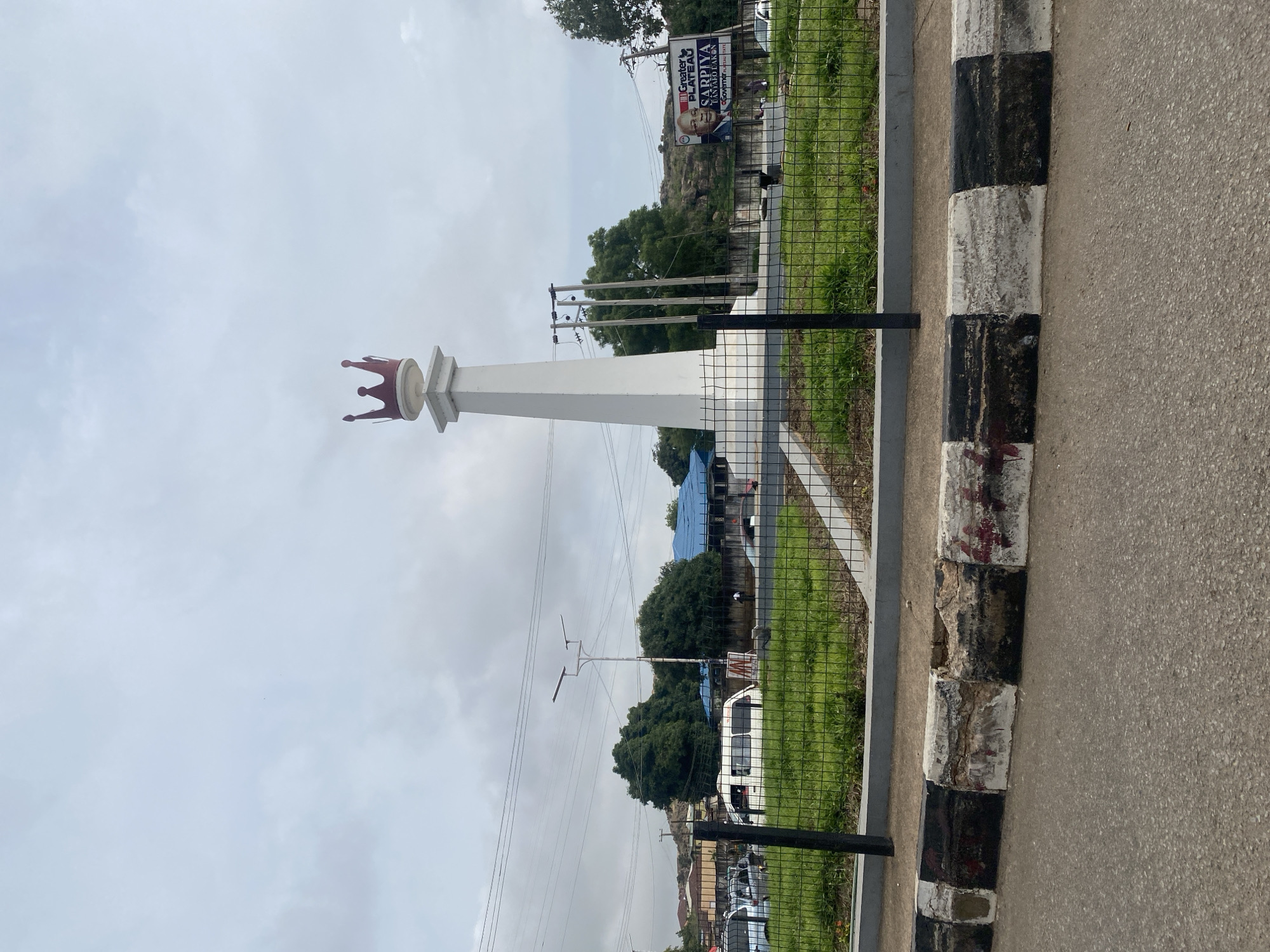 The image, taken in Jos, Nigeria, depicts an outdoor scene dominated by a monument and roadside elements under an overcast sky. A tall, white, obelisk-like monument topped with a distinctive red or maroon crown stands prominently on the left side of the frame.
To the right of the monument, an asphalt road stretches horizontally, bordered by a curb painted in alternating black and white sections, showing visible signs of wear and chipping. A black metal mesh fence runs along the curb, separating the road from a grassy area. This area is populated with green grass and several trees, including a large, leafy tree in the middle ground.
In the background, utility poles with numerous overhead power lines are visible, crisscrossing against the cloudy sky. Beyond these, distant buildings can be seen, one notably featuring a blue roof, indicating a semi-urban or urban environment. A large billboard is situated in the upper right background, displaying the text "GREATER PLATEAU" at the top, followed by "SARPIYA" and "IMAMU LAK," alongside a portrait of a man.
A white van or mini-bus is parked on the grassy area behind the fence, and other vehicles are faintly discernible further down the road. The soft, diffuse lighting suggests it is daytime, likely under an overcast or cloudy sky. The scene captures a public thoroughfare or central point, marked by the crowned monument, possibly a landmark within the city.