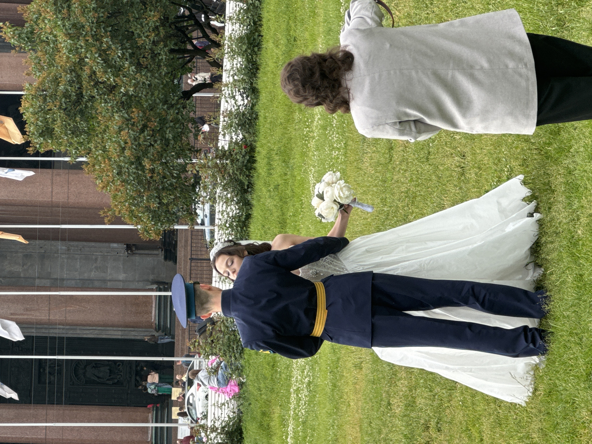 This image captures a wedding scene outdoors on a grassy area in front of a large building with classical architecture in Saint Petersburg, Russia. In the foreground, a newlywed couple is the central focus. The groom, dressed in a dark navy military uniform with a yellow sash, stands with his back to the camera, facing his bride. The bride, wearing a white wedding dress and a veil, leans towards him, their faces close as if sharing a moment. She holds a bouquet of white flowers.
To the right of the couple, a person with curly brown hair is seen from behind, wearing a light gray jacket and black pants. This individual appears to be observing or perhaps photographing the couple.
In the background, the imposing facade of a grand building dominates the scene. It features large columns, a dark entrance with intricate carvings, and flags flying from poles. The architecture suggests a significant public building, possibly a museum, government office, or a historical landmark like St. Isaac's Cathedral, given the architectural style common in Saint Petersburg. The area is lined with manicured bushes and trees, and a white car is visible further in the background along with other pedestrians. The overall atmosphere is that of a special occasion taking place in a well-maintained urban environment.
