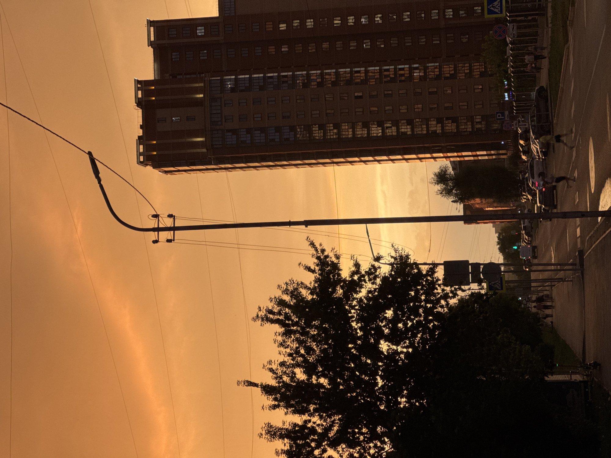 The image depicts a street scene in Novosibirsk, Russia, during a sunset with an orange sky. In the foreground, a sidewalk runs alongside a tree-lined area. People are walking on the sidewalk and crossing a street. The street is lined with cars parked on the side and some driving. A tall, modern apartment building with many windows is visible in the background, illuminated by the setting sun. A street lamp and utility poles with wires are also present. The overall impression is a typical urban evening in Russia.