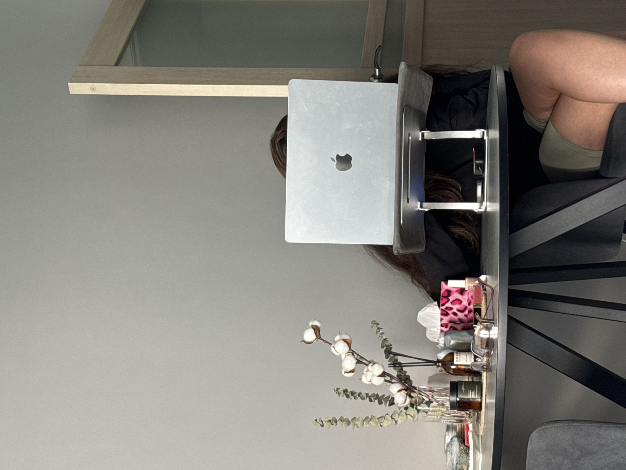 A woman is seated at a desk with a laptop. The desk also has a decorative floral arrangement and various small items. The woman is wearing shorts and has her legs crossed. A framed piece of artwork is on the wall. The location is undetermined, but the mention of Krasnoyarsk, Russia, suggests this is the origin of the image, though the scene itself provides no specific geographical markers.