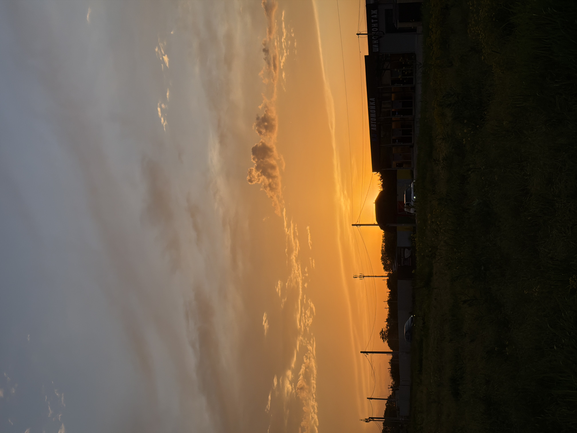The image shows a sunset in Krasnodar, Russia. The sky is predominantly orange, with clouds visible. A building with text "МОЙ АВТОМОЙКА" and "ШИНОМОНТАЖ" is on the right side of the image. Power lines are visible, and there is tall grass/foliage. A car is parked next to the building.