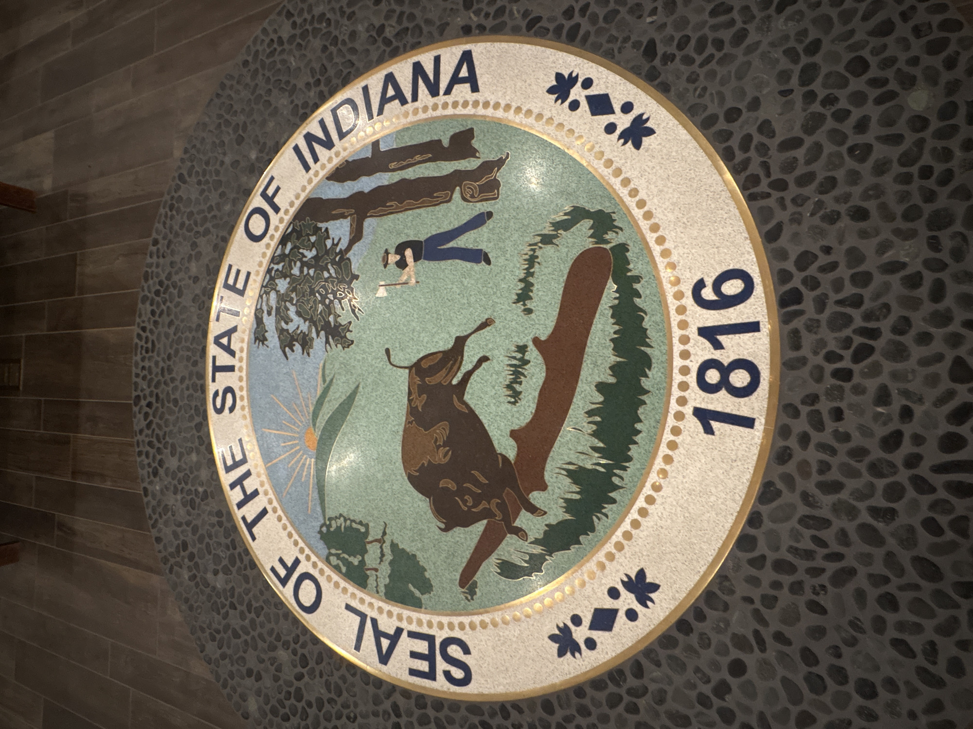 The image shows a circular seal embedded in a floor made of small, dark gray stones. The seal is the "Seal of the State of Indiana" and features a central design of a bison jumping over a log, with a frontiersman chopping down a tree in the background. A sunrise with rays is visible above rolling hills. The text "SEAL OF THE STATE OF INDIANA" encircles the upper portion of the seal, and the year "1816" is prominently displayed at the bottom. The seal is bordered by a thin gold ring and adorned with blue floral or star-like emblems on either side. The flooring around the seal is consistent with the surrounding dark stone. The image does not provide any specific information or visual cues that indicate its location within New Harmony, United States.