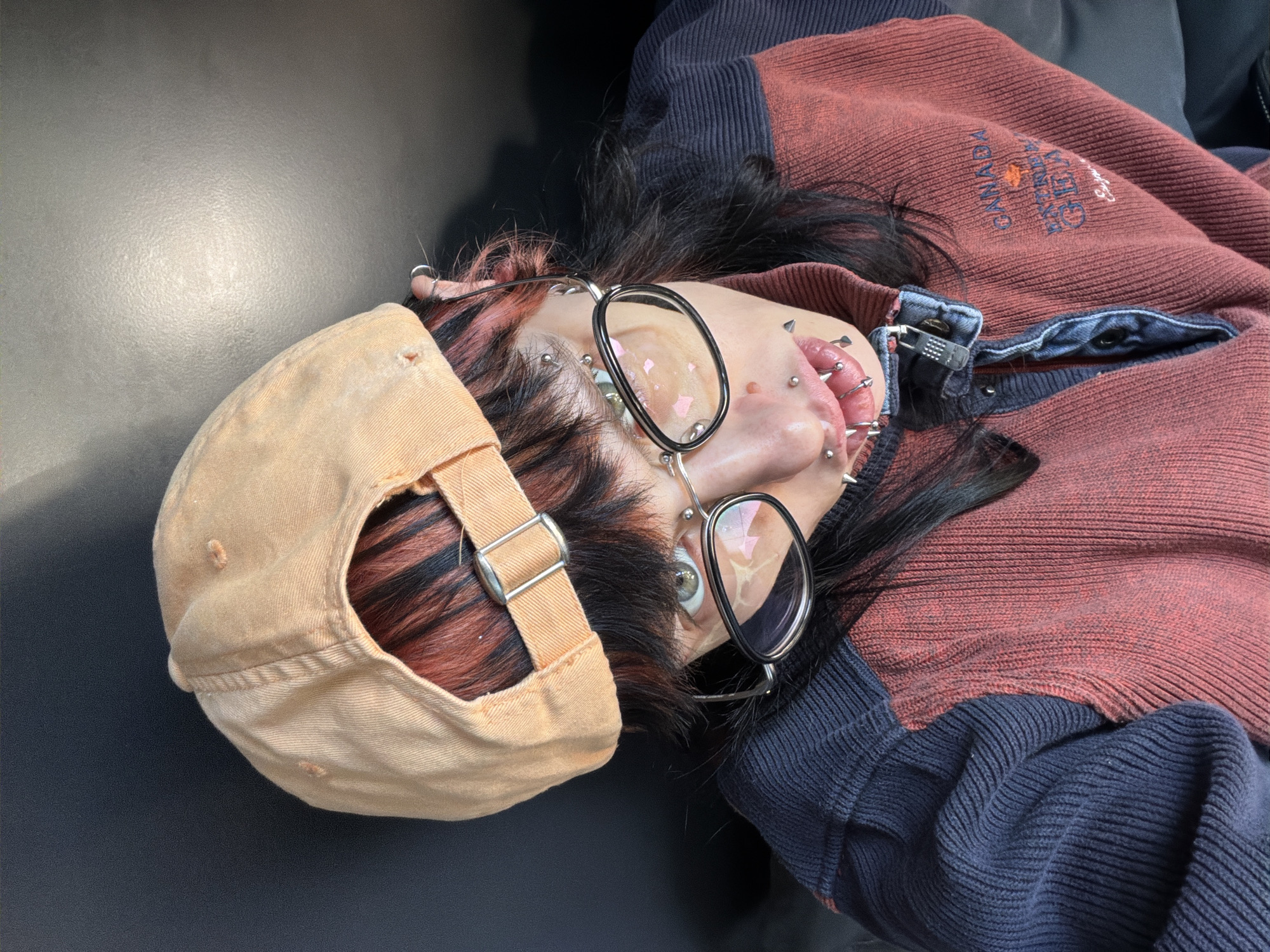 An individual lies on a dark surface, possibly black fabric or leather. They are wearing a tan baseball cap with a silver buckle on the back strap, black hair with red streaks peeking out from under the cap. The person has numerous piercings on their face, including around the lips, nose, and ears. They are wearing round, thin-rimmed glasses. The person is dressed in a sweater, reddish-brown on top with the word "CANADA" and another word that is unclear embroidered near the chest, and navy blue knit on the bottom. The location is not evident in the image, so it can not be determined from the image if it is in the city of Busmantsi, Bulgaria.