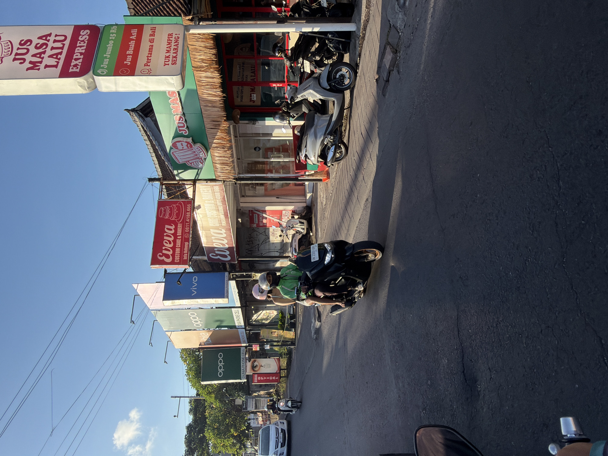 This image captures a vibrant street scene in North Kuta, Indonesia, bathed in bright sunlight with long shadows, suggesting either morning or late afternoon. The sky is clear blue with a few scattered clouds.
The scene depicts a bustling commercial street lined with various shops and businesses. Several motorcycles and scooters are visible; some are parked along the sidewalk, while others are on the asphalt road, including one with a driver and passenger. The driver wears a green jacket and a white helmet, and the passenger wears a pink helmet. A white car is also visible further down the road. Power lines crisscross the upper part of the frame, connecting to utility poles. Buildings feature distinctive thatched or straw awnings, adding to the local architectural style.
Numerous signs are prominently displayed along the street. Key text includes:
- A large sign for "JUS MASA LALU EXPRESS" advertising "Jus Jumbo 15 Rb" (Jumbo Juice 15,000 IDR), "Jus Buah Asli" (Original Fruit Juice), and proudly stating "Pertama di Bali" (First in Bali) with the call to action "YUK MAMPIR SEKARANG!" (COME VISIT NOW!). Another sign nearby plays on words with "MANTAN MARI CIPTA KENANGAN BARU SAMA JMAA LATI" (Ex, let's create new memories with JMAA LATI).
- Multiple signs for "Eveva CUSTOM CAKE & BAKERY" with contact information "Info Hubungi 0811.4188.666".
- Brand advertisements for "vivo" and "oppo," alongside signs for "OLMA PHONE CELL," "DUNIA PHONE CELL," and "BUANA OPTICAL."
- A motorcycle license plate "DK 2301 UBI," indicating registration in Bali.
The image portrays a typical daily street activity in a lively urban area in Bali, characterized by local businesses and abundant motor vehicle traffic.