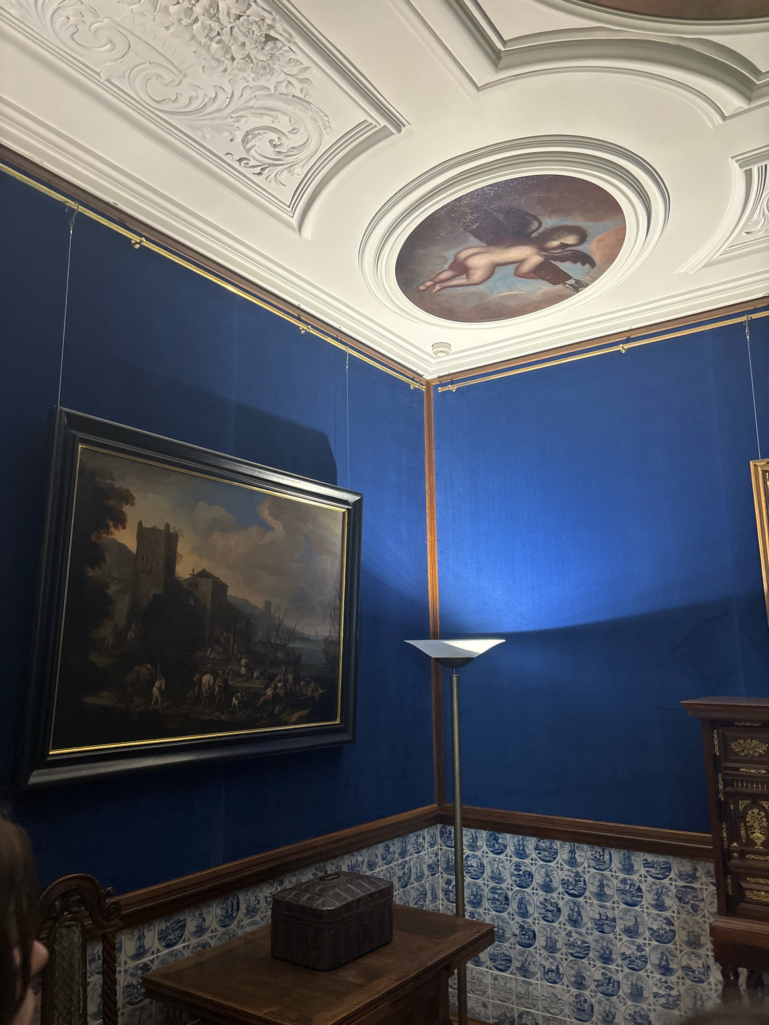 This image shows a richly decorated interior room, likely within the Hermitage Museum in Saint Petersburg, Russia. The walls are covered in a vibrant, deep blue fabric, creating a striking contrast with the white ornamental plasterwork on the ceiling. The ceiling features a central oval painting of a winged cherub in a classical style, surrounded by intricate floral and scroll motifs.
A large, dark-framed painting depicting a harbor scene with a castle and numerous figures is hung on the left wall. The painting is suspended from the ceiling by thin wires. To the right, a modern floor lamp with a triangular shade casts a pool of light onto the blue wall.
Below the paintings, the lower portion of the walls is adorned with blue and white tiles featuring maritime scenes with ships. A dark wooden table with a decorative chest sits in the foreground. Another ornate wooden cabinet is visible on the far right. The overall impression is one of historical opulence and artistic display.
