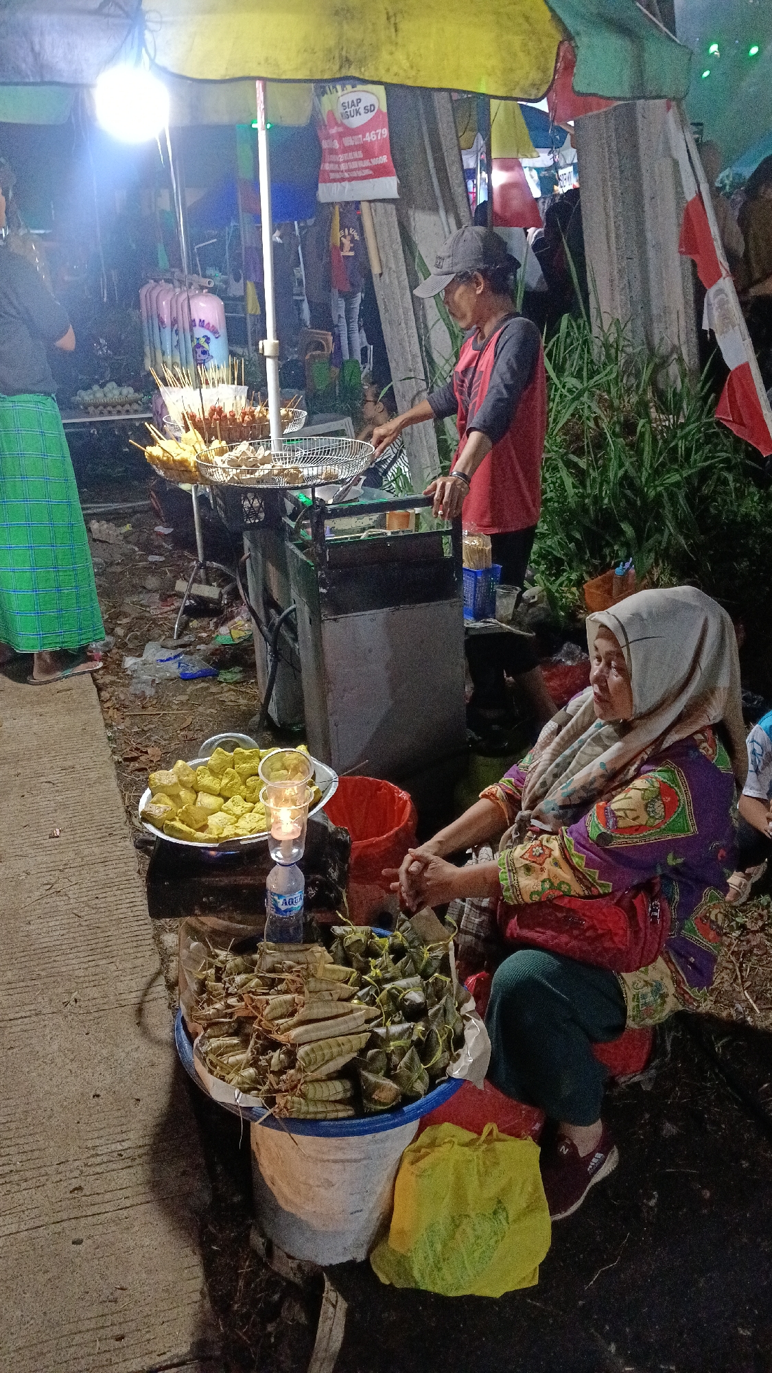 At night in Tajurhalang, Indonesia, a street food vendor with a headscarf and a patterned purple, yellow, and green tunic sits behind a display of rice cakes wrapped in leaves. Next to her, on a wooden surface, is a tray of fried tofu, a red bucket, and a bottle of Aqua water with a candle holder made of stacked plastic cups. In the background, a younger vendor in a red and black shirt and a black baseball cap is preparing food, possibly frying it, over a stove. Behind them, colorful awnings and flags are visible, along with tall green plants and a concrete wall. A woman in a green checkered skirt stands to the left, partially visible. The scene is illuminated by a bright overhead light.