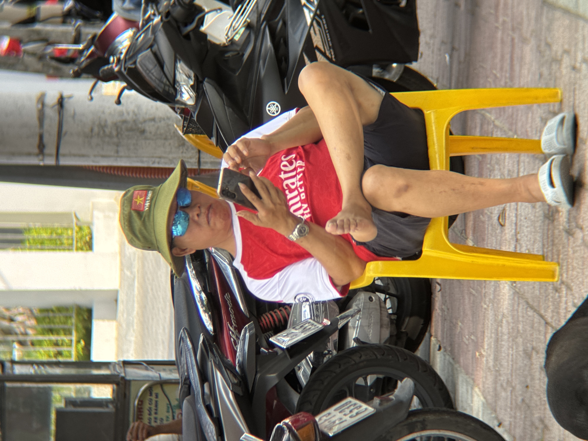 In Nha Trang, Vietnam, a man sits on a yellow plastic chair in front of parked motorcycles. He is wearing a green bucket hat with a red star and "Việt Nam" text, blue mirrored sunglasses, a red and white t-shirt with "Emirates Fly Better" printed on it, and dark shorts. He is holding a smartphone in his right hand and looking at it. His left leg is crossed over his right, and he is barefoot with white slides on his feet. The environment appears to be a street or sidewalk, with the motorcycles suggesting a transportation hub or parking area. A dark, blurry object, possibly a dog, is visible in the lower left corner.