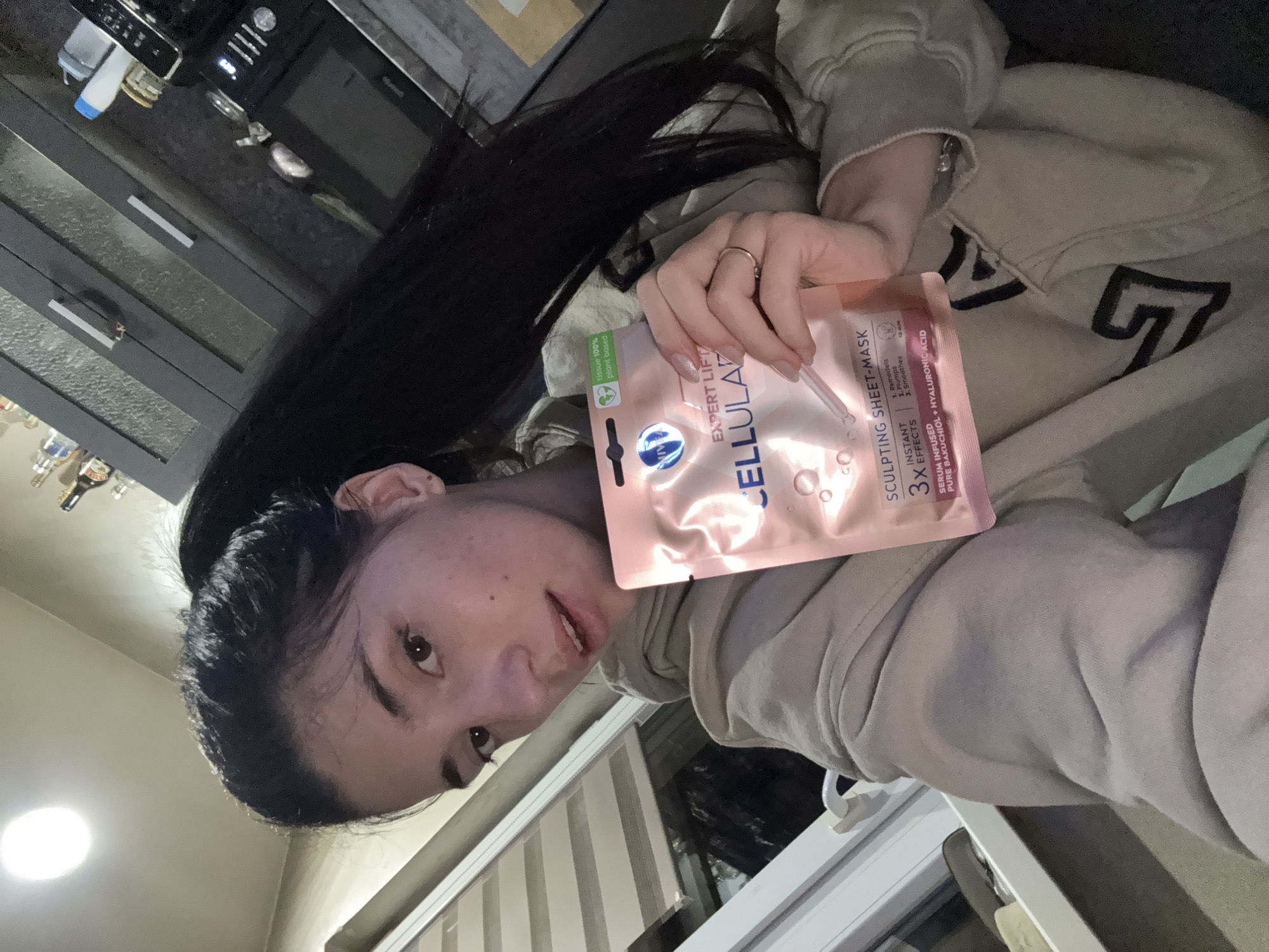 A woman with long dark hair is holding up a package of "Cellular Expert Lift Sculpting Sheet-Mask" in what appears to be an indoor setting. The package is pink with blue and white writing, and it indicates that the mask contains pure bakuchiol and hyaluronic acid. The woman is wearing a light-colored hoodie. A kitchen with dark cabinets and appliances is visible in the background. Slatted blinds are also visible. The location is not discernible as Sofia, Bulgaria, based on the image.