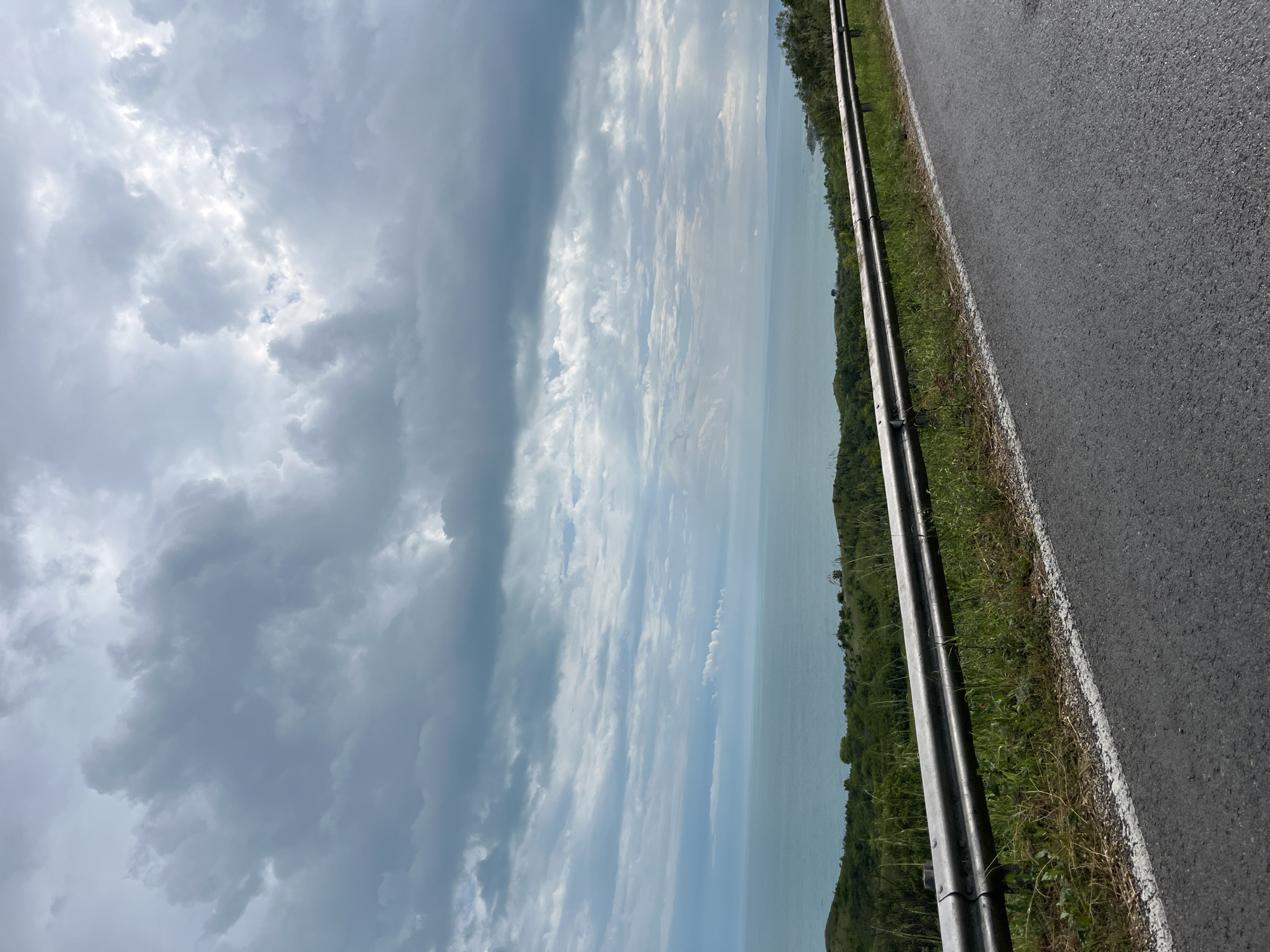 This image presents a wide, elevated view of a coastal landscape in the vicinity of Topola, Bulgaria, under an overcast sky. The upper two-thirds of the frame are dominated by a dense formation of gray and white stratocumulus clouds, exhibiting varying levels of thickness and light penetration, with some brighter areas suggesting diffuse sunlight.
A clear, horizontal horizon line separates the cloudy sky from a calm body of water, consistent with the Black Sea, which appears a uniform blue-green color. Below the horizon, a series of green, undulating hills covered in dense vegetation defines the coastline. Towards the far right, a faint outline of the coastal edge with minor irregularities is discernible.
In the immediate foreground, the edge of an asphalt road is visible, marked by a solid white line. Parallel to the road and situated on a grassy embankment is a two-tier metal guardrail, extending from the lower left to the mid-right of the frame. The grassy verge shows a mix of green and dry vegetation, including scattered wildflowers.
No human figures, animals, vehicles, or specific activities are present in the scene. The overall environment suggests a natural, elevated viewpoint overlooking a coastal region on a heavily cloudy day.