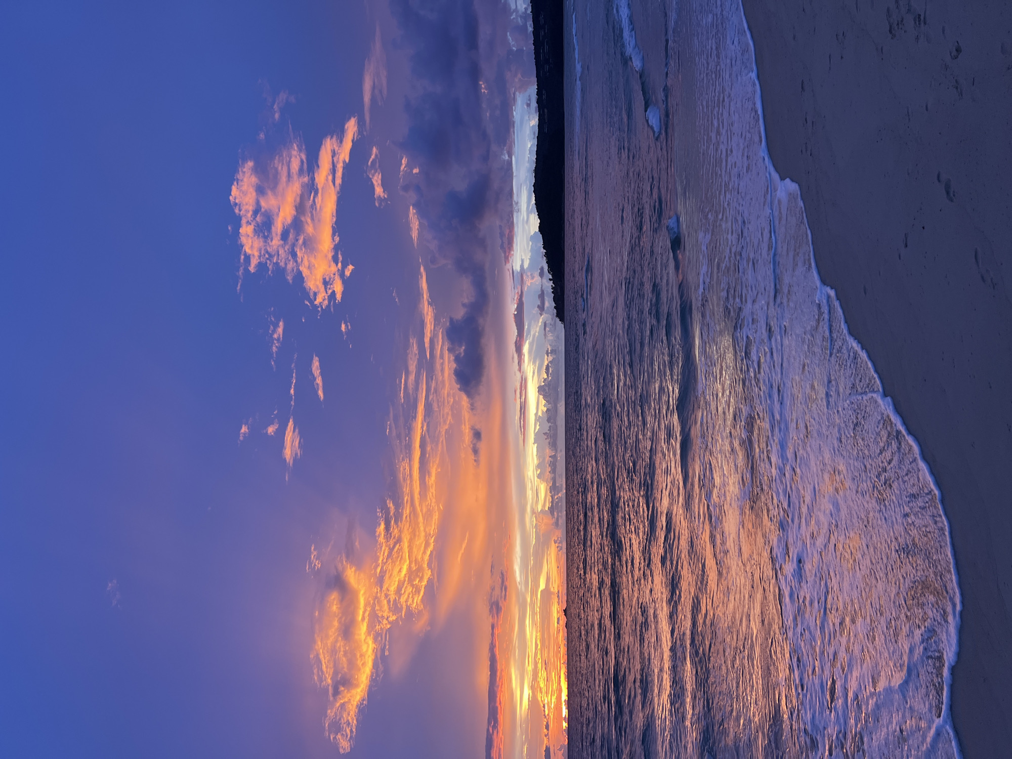 This is a vertical, medium shot of a tropical beach at sunset. The sky is a vibrant display of deep purple and orange, with streaks of light radiating from the horizon. Clouds are scattered across the sky, some tinged with pink and gold from the setting sun, while others are a deep, brooding purple. The ocean is calm, with gentle waves lapping at the shore. The water reflects the colors of the sky, creating a shimmering, iridescent effect. The sand in the foreground is a deep, muted blue, with a few footprints visible. The overall impression is one of peaceful, natural beauty. There are no people or man-made objects visible in the image. The location is not specified.