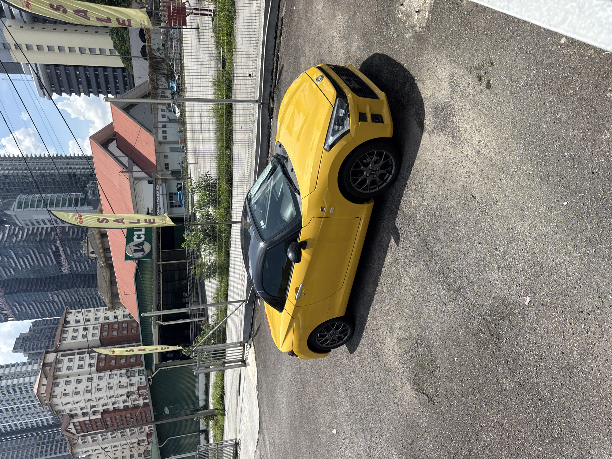 A yellow Daihatsu Copen roadster is parked on a gray asphalt surface, casting a shadow to its right. The car's roof is black. A gray metal fence runs along the left side of the vehicle. Behind the fence, there are green plants and buildings, including a building with a red roof. In the background, there are tall buildings under a sky with some clouds. There is a banner that reads "SALE". The location appears to be an urban area in Kuala Lumpur, Malaysia.