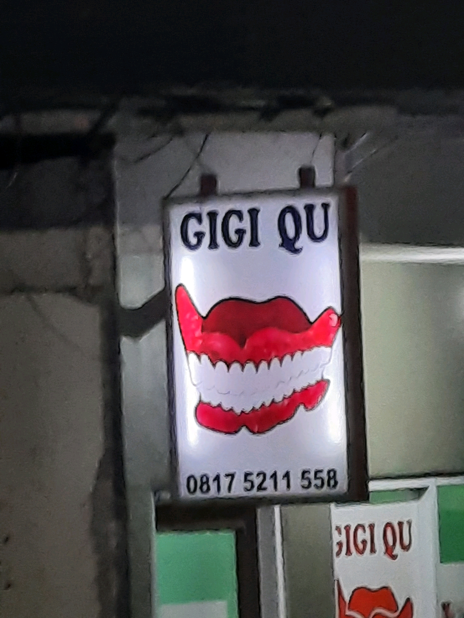 The image shows a lit sign advertising a dental clinic named "GIGI QU" in East Jakarta, Indonesia. The sign features a cartoonish depiction of a mouth with bright red lips and white teeth. Below the graphic, a phone number "0817 5211 558" is displayed. Another smaller sign with the same name and graphic is partially visible to the right. The signs are mounted on a dark, rough-textured wall. The surrounding environment is dimly lit, suggesting the photo was taken at night or in a poorly lit area.