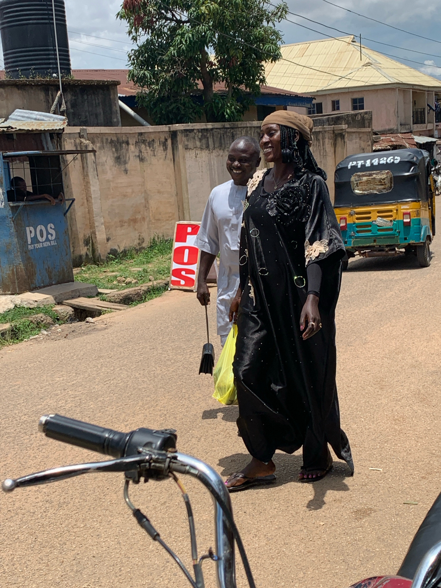 In Jos, Nigeria, on a paved street under a cloudy sky, a man in a white shirt and a woman in a black abaya are walking. The woman, wearing a brown headscarf and sandals, carries a yellow bag and a small broom. The man, also in sandals, smiles and walks beside her. Behind them, a yellow and green auto-rickshaw with license plate PT14265 is parked. To the left, a blue kiosk with "POS PAY YOUR NEPA BILL" signage is visible, with a person leaning out of a window. A wall runs along the street, with trees and buildings in the background. In the foreground, a motorcycle handlebar and brake lever are in sharp focus.