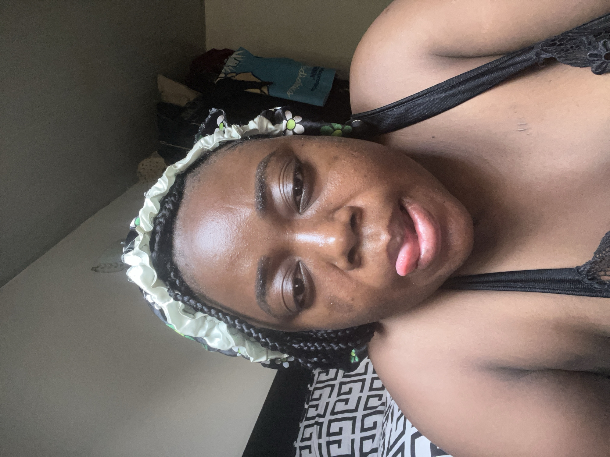 A close-up, rotated selfie captures a person with dark skin, likely female, in a relaxed indoor setting, possibly a bedroom in Abuja, Nigeria. The subject wears a satin-like protective hair bonnet with a light green and white ruffled edge, adorned with a floral or leaf pattern and small green beads, covering braided hair. She looks towards the camera with her tongue slightly extended, conveying a playful or casual expression. Her skin exhibits a natural sheen.
She is wearing a black bra or top with a visible lace trim on the right side of the frame. Below her arm, a black and white geometric patterned fabric, likely bedsheets, is visible. The background consists of a plain, dark-colored wall behind her head, and to the upper left, an out-of-focus area with cluttered items, possibly folded clothes or bedding. Among these items is a light blue fabric with some indistinct white markings or partial text. The lighting is soft and ambient, consistent with an indoor environment. No specific time of day or weather can be determined from the visual cues.