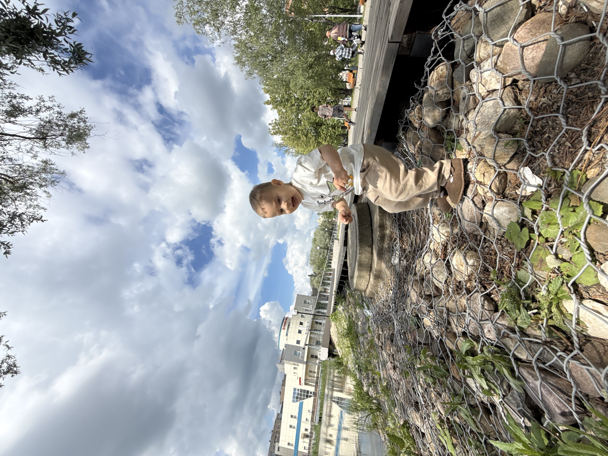 A bright outdoor scene in Rusanovo, Russia, captures a joyful young child next to a gabion wall. The child, wearing a white t-shirt with a graphic, light brown pants, and shoes, is positioned in the foreground, looking towards the camera with an open mouth, conveying happiness or excitement.
To the child's right and extending into the foreground is a gabion wall, a structure of metal mesh filled with rocks, with some green plants growing through and around it. Behind the child and to the left, a wooden boardwalk or path is visible, where several blurred adult figures can be seen walking or standing. Lush green trees line the boardwalk in the mid-ground.
In the distance, under a sky with prominent white cumulus clouds and clear blue patches, a cityscape of modern multi-story buildings is visible. One building prominently features a red circular logo with white text, which clearly reads "Аптека" (Russian for Pharmacy). The image depicts a sunny day with scattered clouds, suggesting a pleasant atmosphere in a park or waterfront setting.
