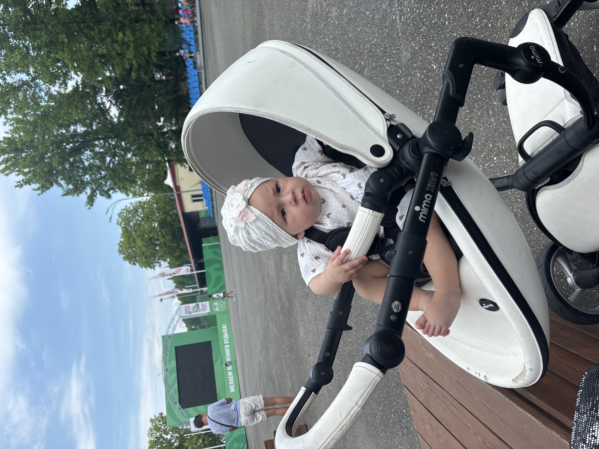 In Sochi, Russia, on a cloudy day, a baby sits in a white stroller. The stroller's handle bars are black, and the brand name "mima xari" is visible. The baby wears a white turban with a pink flower and a light-colored shirt with heart patterns. The background features a green stage with a large screen, trees, a seating area with blue benches, and asphalt ground. A person in a blue and white patterned shirt and white shorts stands in the distance, facing away from the camera, near the stage. Other people are also visible in the background, some sitting on the benches.