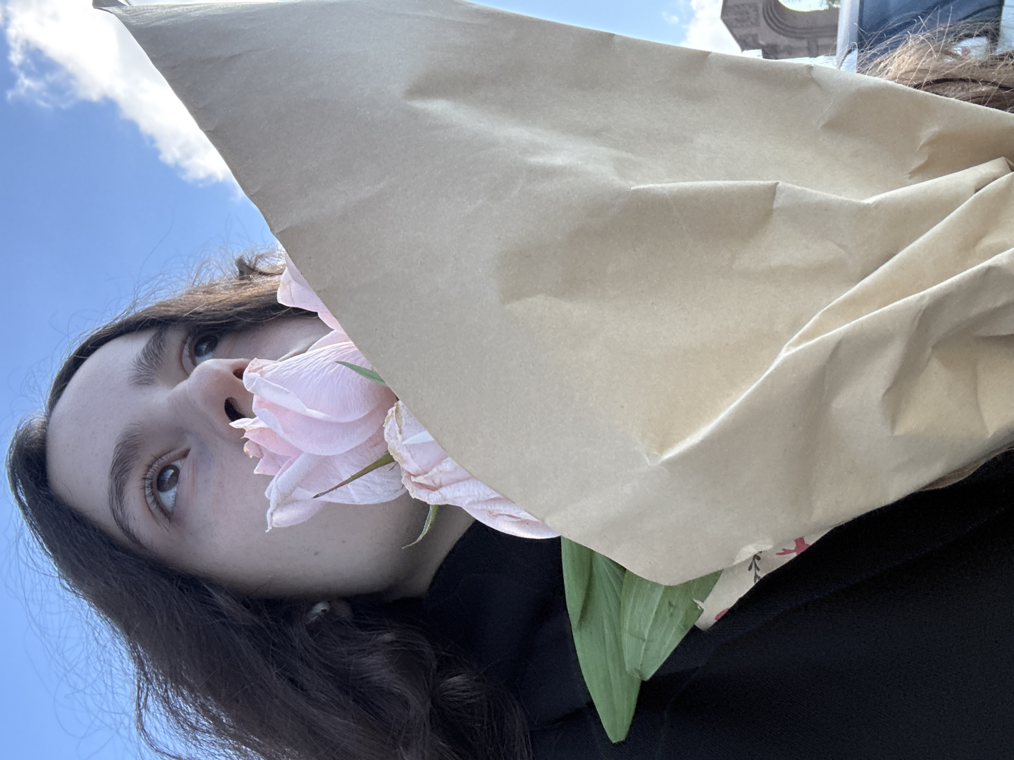 A low-angle shot depicts a person holding a bouquet of pink roses in a brown paper bag. The person has dark hair and is wearing a black shirt or jacket. The background shows a blue sky with some clouds. A building with architectural details is faintly visible in the top right corner. This photograph was taken in Yerevan, Armenia.