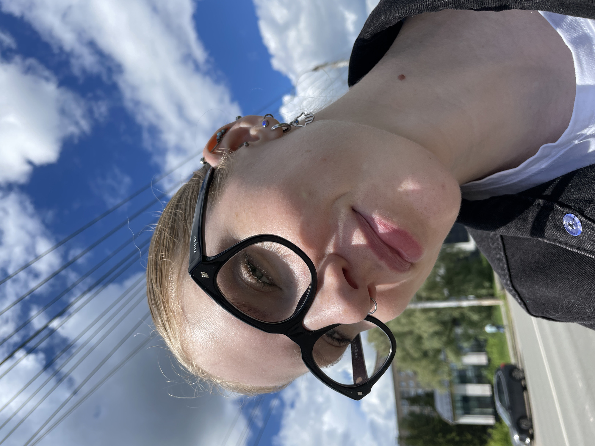 A low-angle, slightly rotated, close-up photograph shows a person wearing black Vogue glasses and various ear piercings. The person has light skin and blonde hair. They also have a small nose ring. They are wearing a white shirt underneath a dark jacket with a visible blue button. In the background, there is a blue sky with white clouds and architectural structures, including a bridge or a building with support cables. The presence of buildings and vehicles indicates an urban environment. Based on the information available, the location is Izhevsk, Russia.