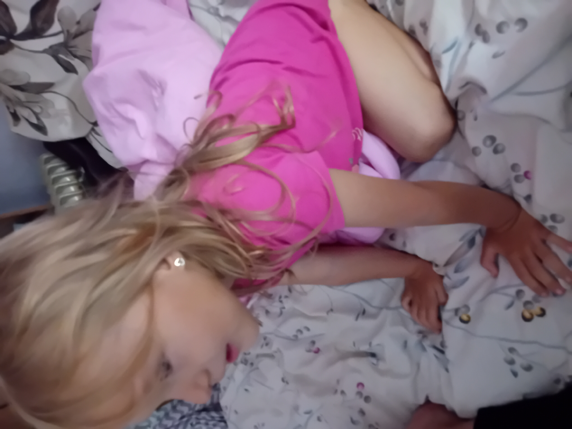 The image shows a young girl lying on a bed. She has blonde hair and is wearing a pink shirt. The bed is covered in a floral patterned sheet. The girl appears to be relaxed. The lighting suggests it is daytime. The location is Chuhuiv, Ukraine. There is no visible text in the image.