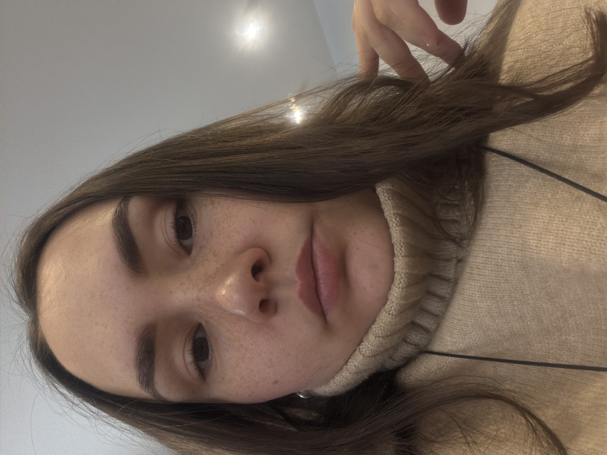 A selfie of a fair-skinned young woman with brown hair and freckles is captured indoors. She is wearing a beige turtleneck sweater. Her right hand is partially visible near the top of the frame, touching her hair. The image is taken at an angle, with a ceiling light visible in the background. The specific location in the city of Chaikovsky, Russia cannot be determined from the provided image.