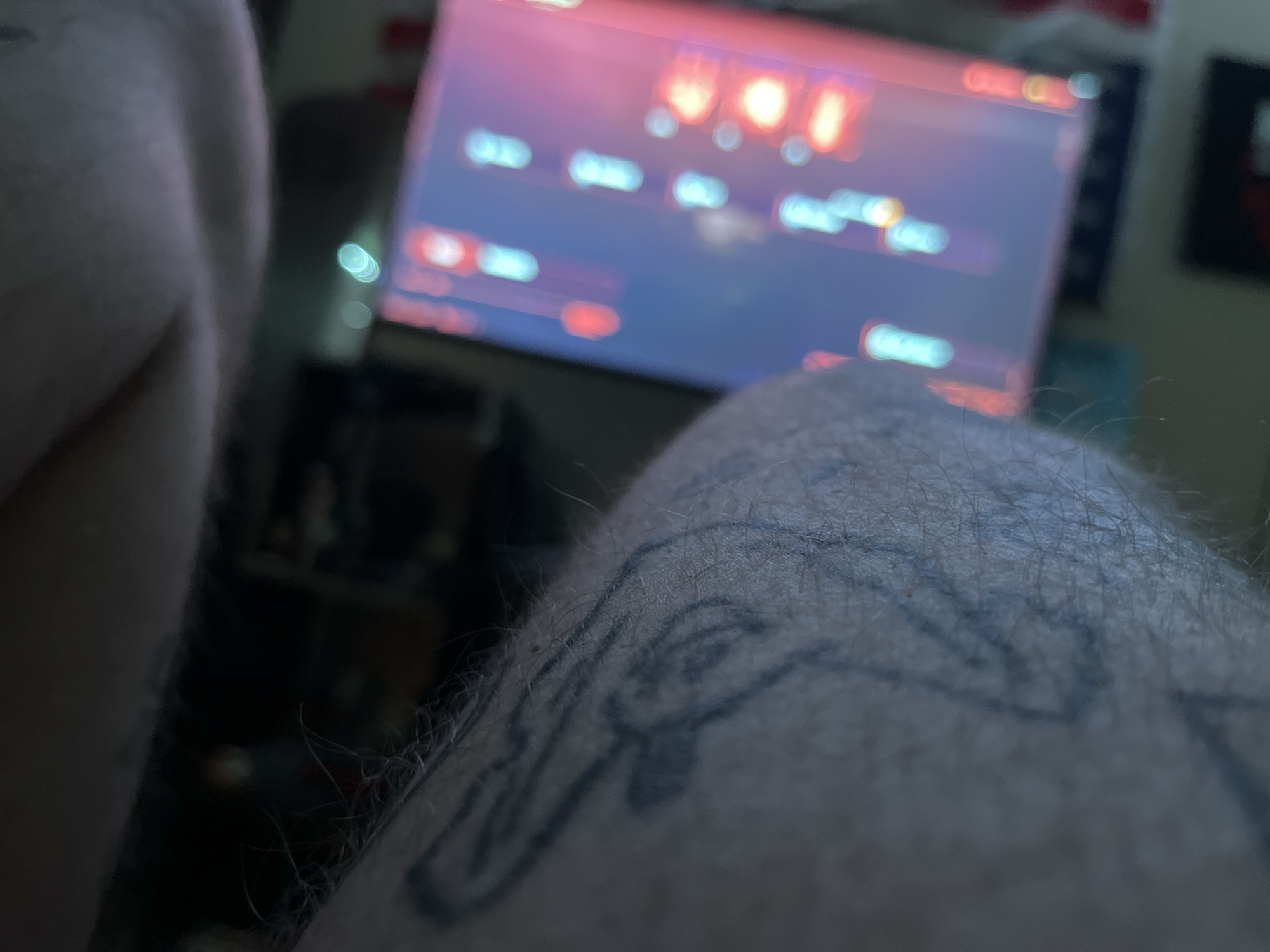 The image is a close-up, out-of-focus shot of a person's leg with a tattoo. In the background, a computer monitor displays a colorful, blurry interface. The lighting is dim, with the monitor being the primary light source. The scene appears to be indoors, likely in a home environment. The subject is not clearly identifiable due to the focus and framing.
