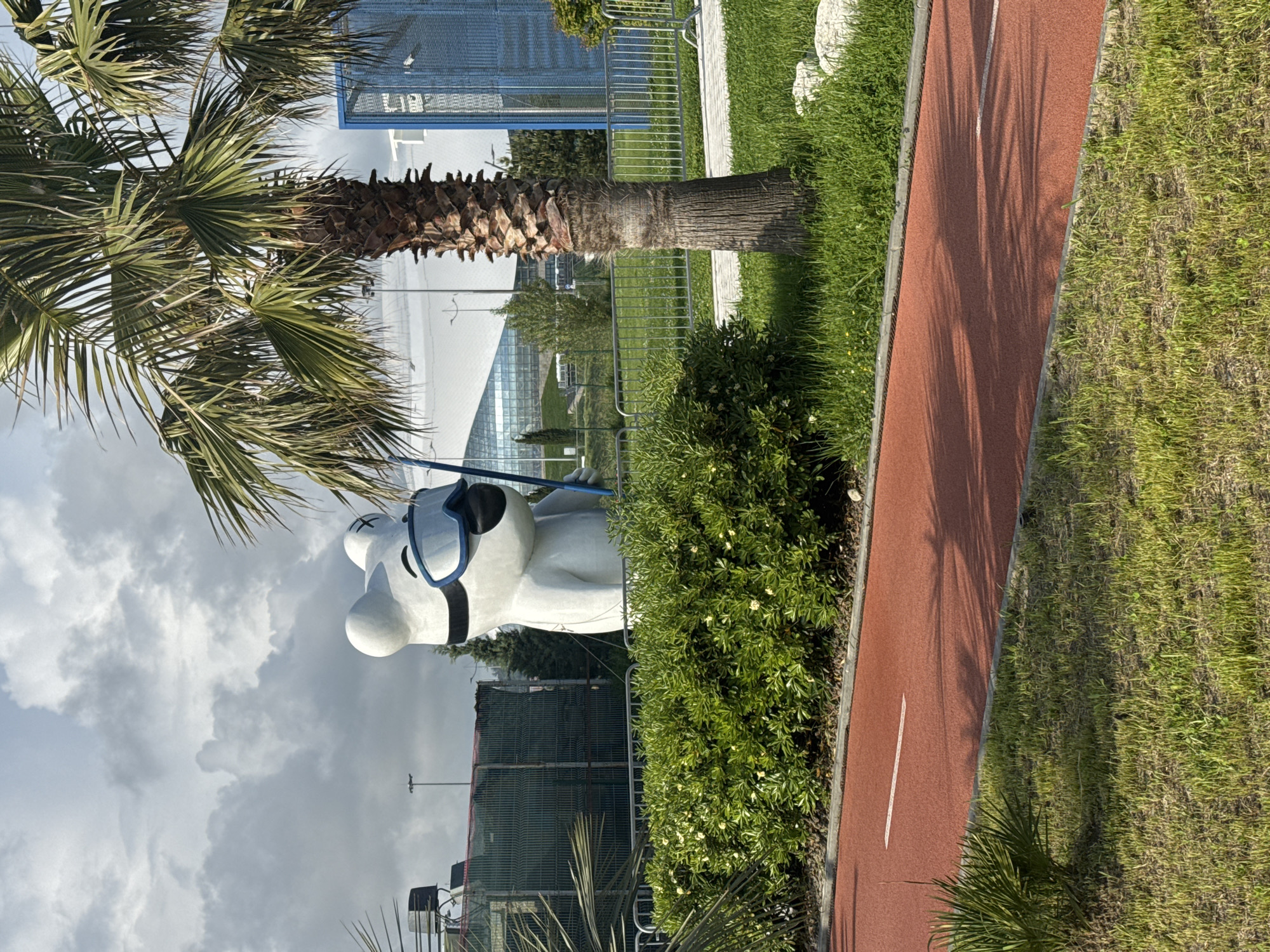 The image depicts a scene in Sirius, Russia, featuring a whimsical sculpture of a white bear wearing blue-rimmed sunglasses. The sculpture stands beside a palm tree and lush green shrubbery, bordering a red running track. In the background, a modern glass-paneled structure can be seen, partially obscured by fencing and vegetation. The sky is overcast with gray clouds. The scene is generally composed of elements associated with a park or recreational area.
