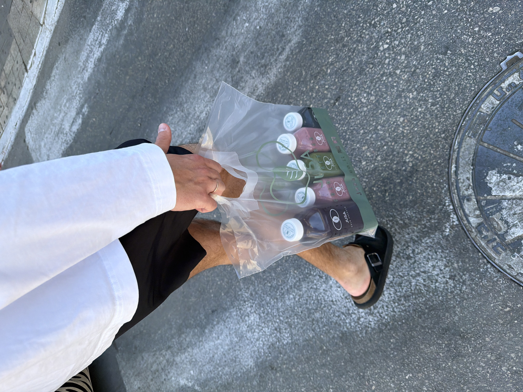 The image, captured from a high-angle overhead perspective, shows a person carrying a bag of bottled beverages on an outdoor paved surface. The subject is partially visible, wearing a white long-sleeved top, black shorts, and a black Birkenstock-style sandal on their right foot. A gold ring is worn on the person's left ring finger.
They are holding a clear plastic bag containing at least six bottled drinks. The bottles are distinct colors: some appear pink or purple, others are green, and some are a dark brown or black. The plastic bag features a green section with white text that clearly reads "JUSA" and "COLD PRESSED," with additional smaller text "JUSA go fit!". The white caps of the bottles also display the "JUSA" brand name and a stylized leaf logo, which is repeated on the bottle labels.
The setting is an outdoor street or sidewalk, characterized by a textured gray asphalt or concrete surface. A metal manhole cover, partially visible on the right side of the frame, shows wear and some indistinct markings. Bright daylight illuminates the scene, suggesting a sunny daytime environment. The image depicts a casual moment, likely of someone walking or briefly pausing with their purchased drinks in Tel-Aviv, Israel.