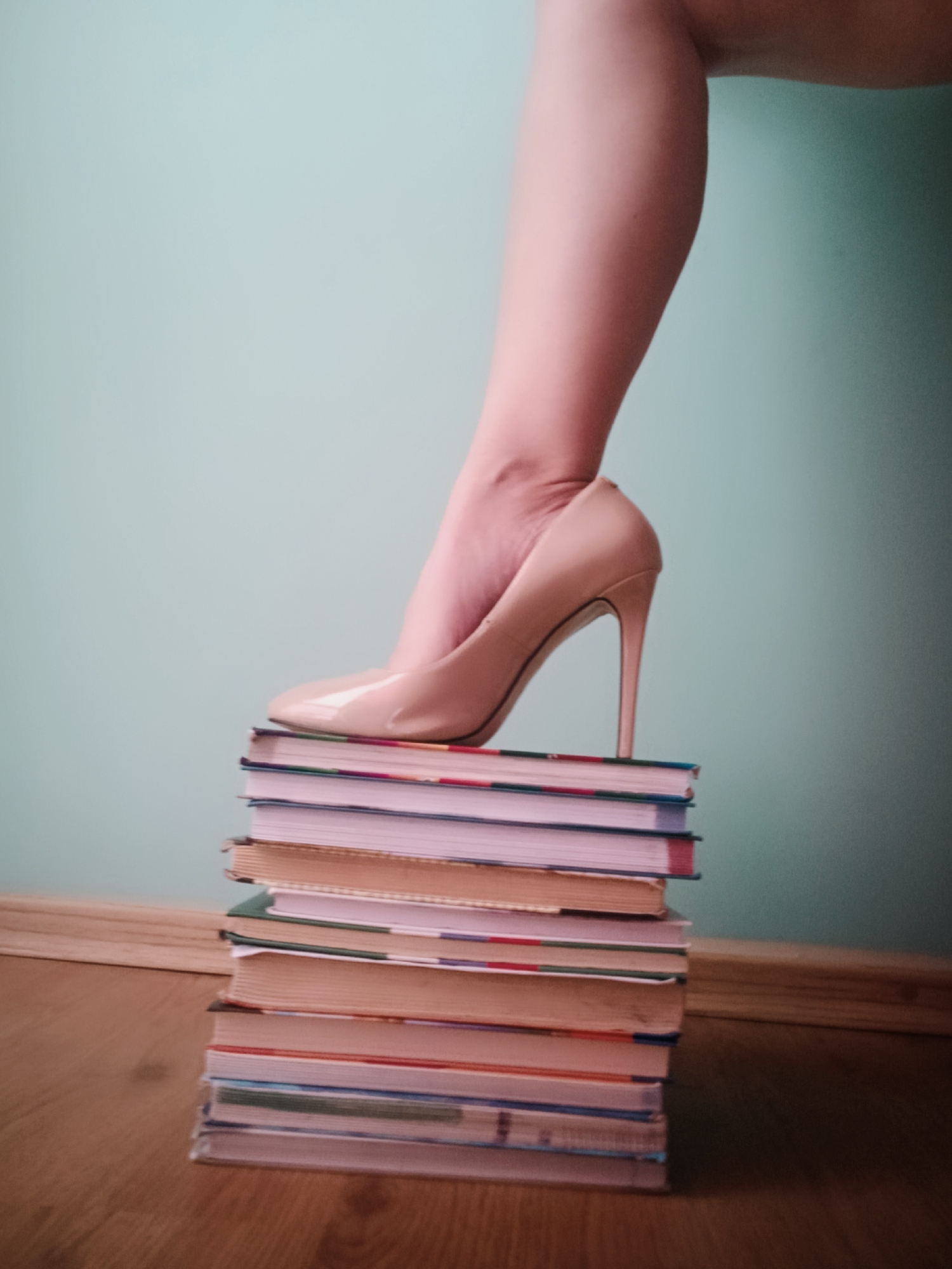 A medium, indoor shot depicts a bare leg with a foot in a nude patent leather high heel shoe resting atop a stack of books. The stack of books is situated on a wooden floor. The background is a muted blue-green wall. The setting appears to be indoors. The image does not provide enough information to determine if the location is Chaikovsky, Russia.