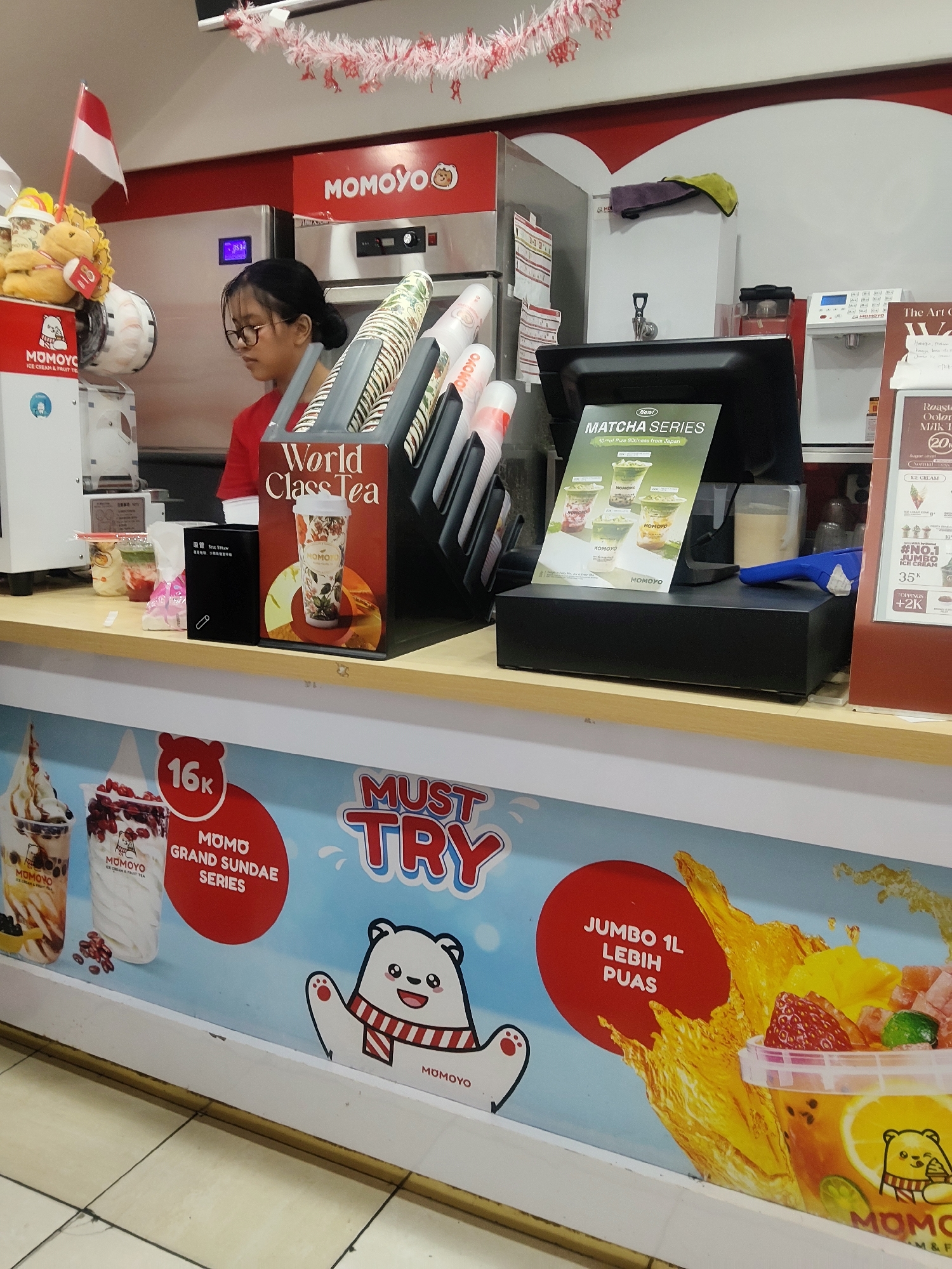 A young woman with dark hair and glasses, wearing a red shirt, stands behind a counter in what appears to be a food service establishment, possibly a boba tea shop. She is looking down, likely at her work. The counter is filled with various items, including cups, a display for "World Class Tea," a menu for "Matcha Series," and a cash register. In the background, there are red and white festive decorations, suggesting a celebration or national holiday, possibly Indonesian Independence Day given the Indonesian flag visible on the left. The establishment's branding, "MOMOYO," is prominent on signage and products. A large advertisement on the lower part of the counter depicts various beverages and promotions, including a "JUMBO 1L LEBIH PUAS" (Jumbo 1L more satisfying) offer and images of ice cream and drinks. The floor is tiled with a light beige and brown pattern. The overall scene depicts a commercial setting with a focus on beverage sales.