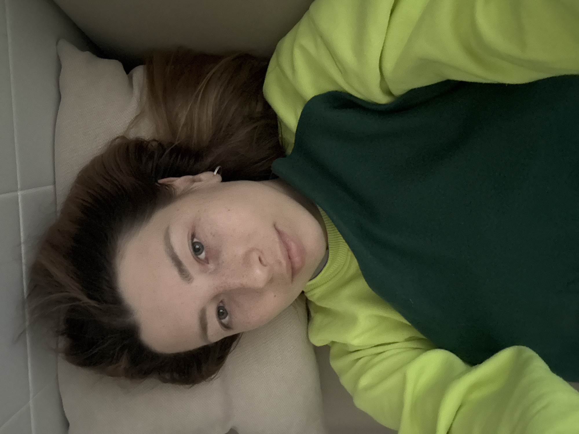 A fair-skinned person with light brown hair is lying on a white pillow, wearing a green and yellow sweatshirt. The background suggests a residential interior. The location cannot be determined from the image.