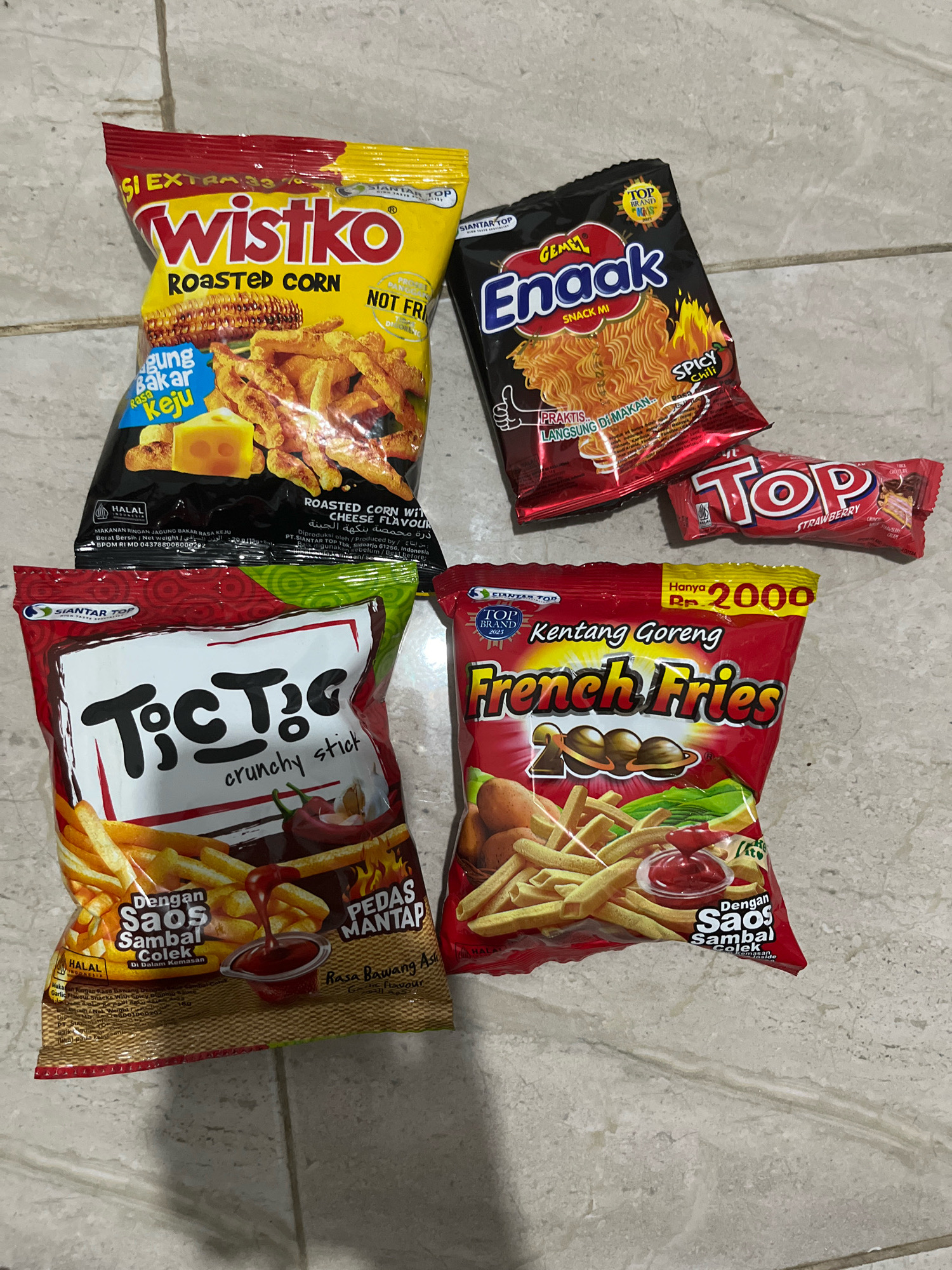 The image displays a flat lay of various snack food packages on a tiled surface. The snacks are arranged in a semi-scattered manner. In the top left is a yellow and red bag labeled "Wistko Roasted Corn" with an image of corn and cheese. To its right, a black bag with red and yellow accents reads "Guel Enaak Snack Mi" and features a depiction of instant noodles with flames. Below the "Wistko" bag and slightly to the left is a red bag with green accents labeled "TiCTiC crunchy stick" showing french fries and chili peppers. To the right of the "TiCTiC" bag is a bright red bag of "Kentang Goreng French Fries" with an image of french fries, potatoes, and ketchup. A small red wrapper with "TOP Strawberry" text is positioned between the "Guel Enaak" and "Kentang Goreng" bags. The scene is lit from above, casting some shadows. The floor appears to be made of light-colored ceramic tiles with visible grout lines. The image is taken from a top-down perspective. The location is identified as Matesih, Indonesia.