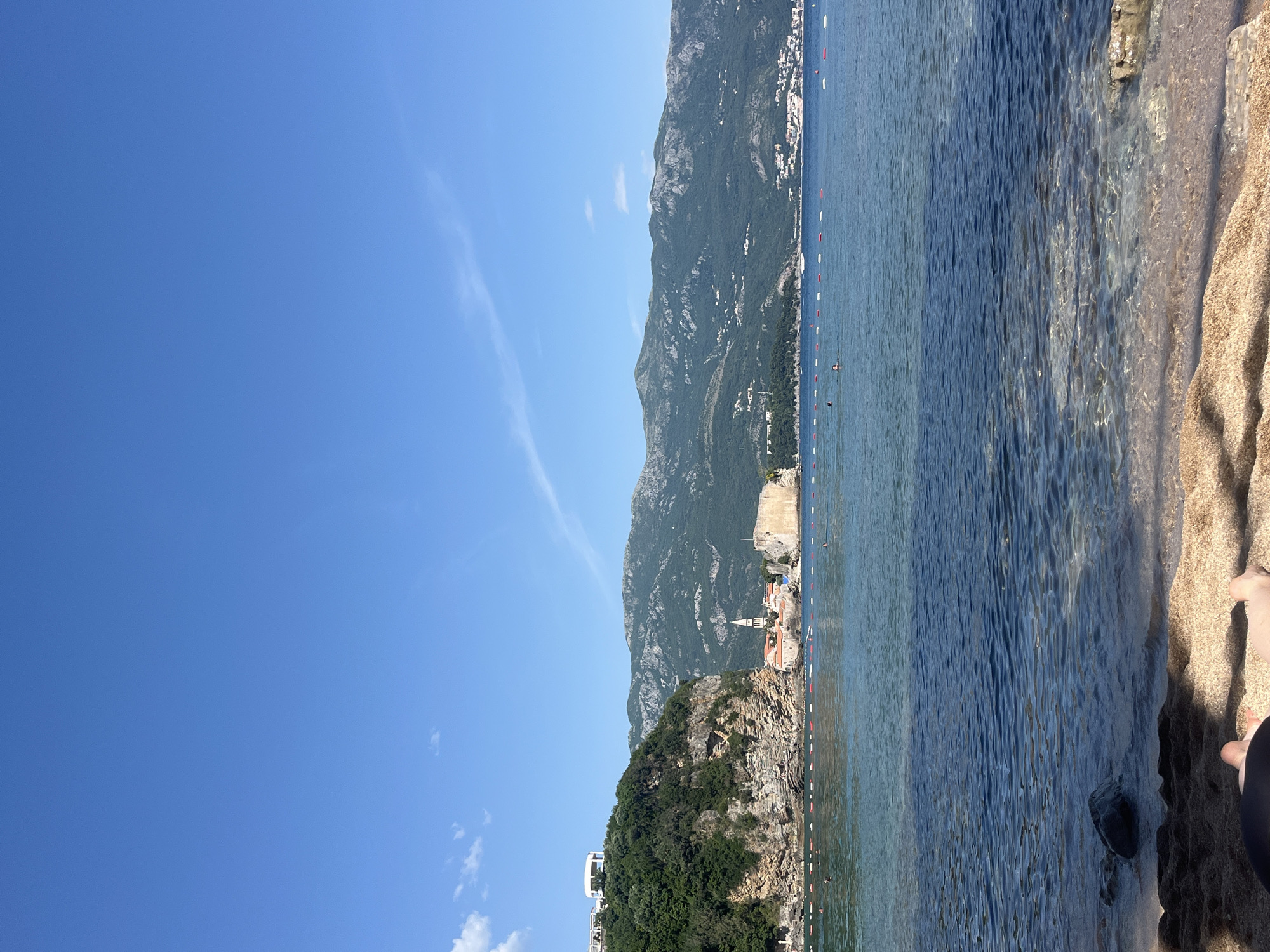 The image shows a coastal view in Budva, Montenegro. The scene includes a clear blue sky with a few faint clouds, a mountain range, a peninsula with dense green vegetation, and the Adriatic Sea. The historic Old Town of Budva is visible on the shoreline. The sea transitions from clear blue to a lighter, translucent shade near the sandy beach. In the foreground, a portion of the sandy beach is visible, with water gently lapping at the shore. Part of a human body is on the beach on the right side of the image. The image is oriented vertically.