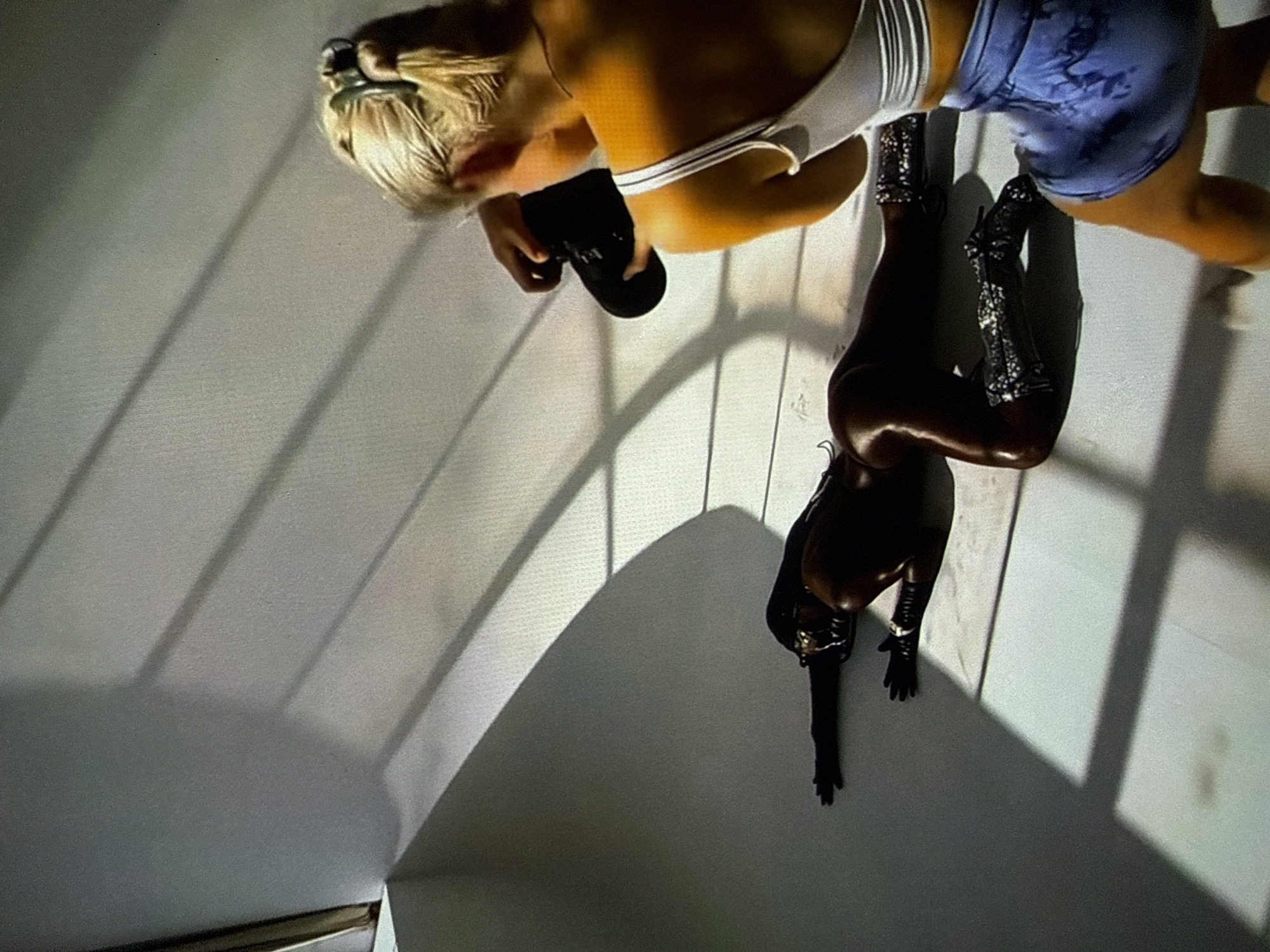 The scene depicts a professional photography session occurring indoors in Varna, Bulgaria. In the center-left foreground, a model with dark skin or heavily shadowed features lies prone on a light-colored floor. The model is positioned on her stomach, propped on her knees and forearms, with her left arm extended straight to the side. She wears a dark, glistening, form-fitting garment, dark gloves, and tall, reflective, possibly silver-sequined boots. Her body surface appears oiled or reflective. In the mid-ground right, a blonde-haired individual, identified as the photographer, is seen from behind, holding a black digital camera. She is wearing a white spaghetti-strap tank top and blue tie-dye athletic shorts, and a thin necklace is visible. The photographer is actively composing a shot, facing the model. The environment consists of plain, light-colored walls and flooring, characteristic of a studio setting. Sharp, defined shadows, appearing as diagonal lines and curved shapes, are cast across the walls and floor, indicating strong, directional lighting. A vertical, narrow, golden-colored pole or column is faintly visible in the far-left background.