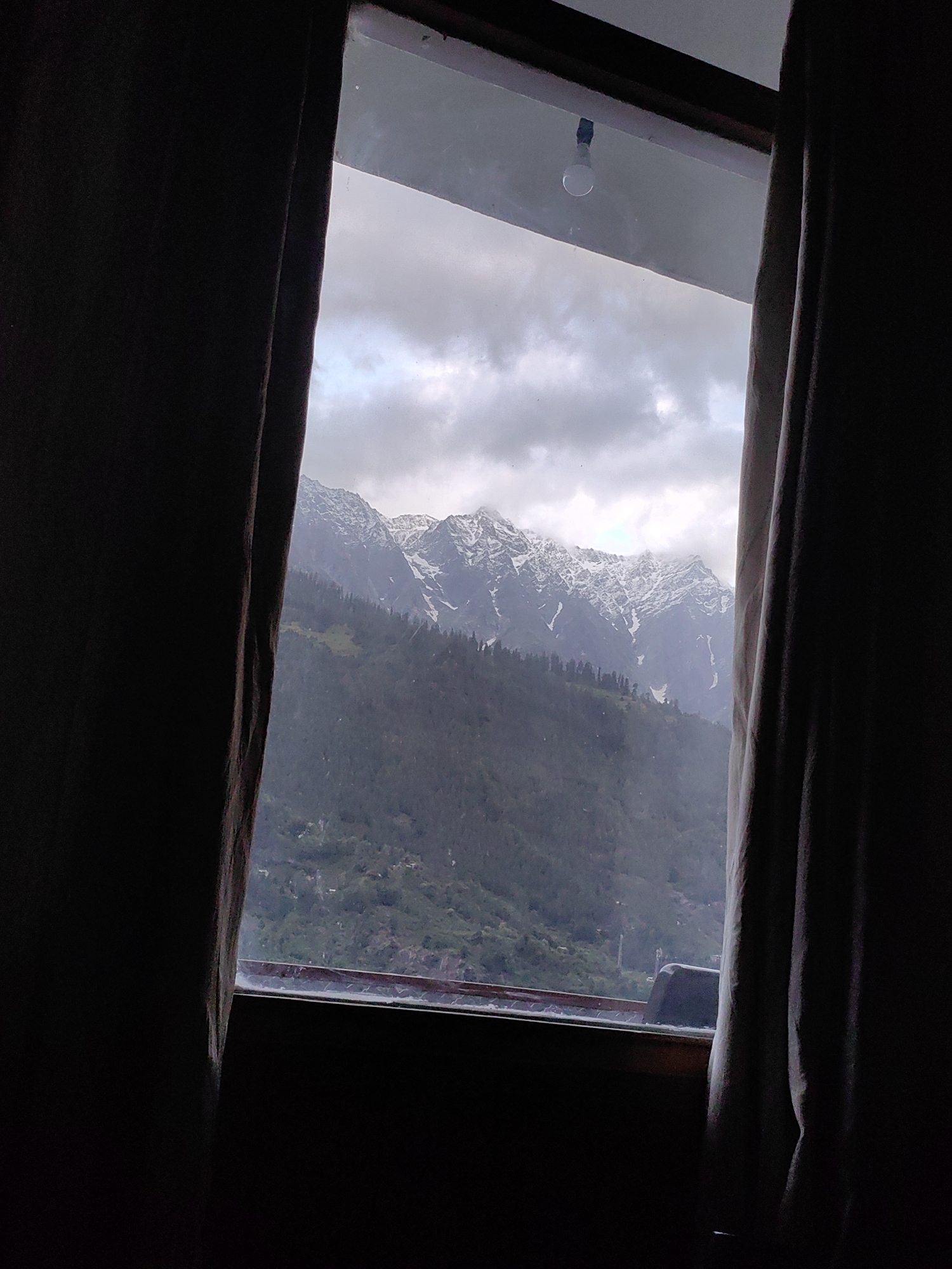 A view through a window in Vashisht, India, reveals snow-capped mountains shrouded in mist and clouds. The foreground is dominated by dark, heavy curtains framing the window. A single, bare light bulb hangs from the ceiling near the top of the window frame. The mountains are covered with a mix of evergreen trees and snow, and the lower slopes show signs of human habitation, with scattered buildings visible through the haze. The overall mood is somber and atmospheric, likely due to the cloudy weather and the limited light.