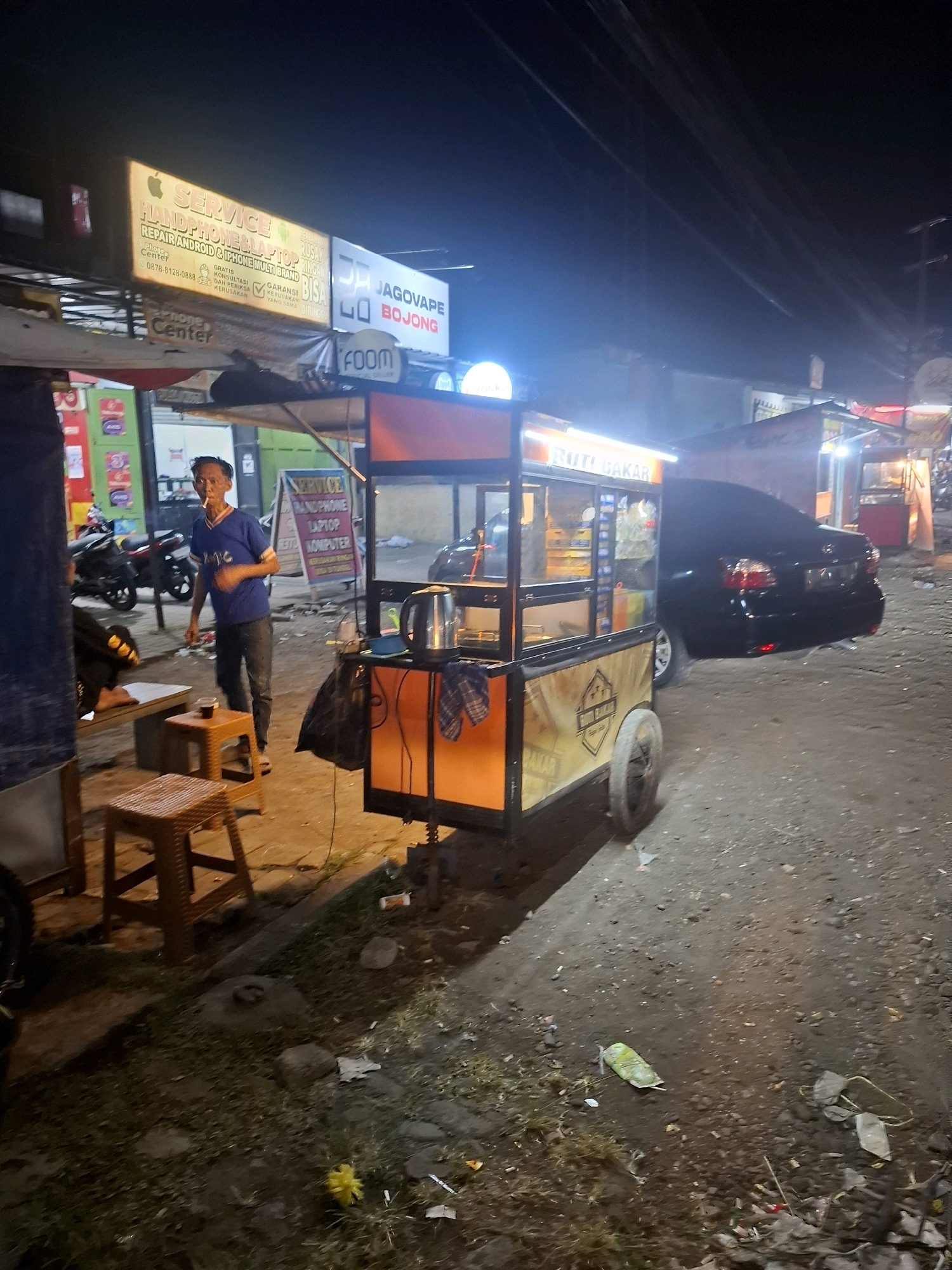 A man is standing next to a food cart at night, in what appears to be a city street in Muka, Indonesia. The food cart, which is orange and yellow, has a sign on it that reads "ROTI BAKAR," which translates to "grilled bread." The man, wearing a blue shirt and dark pants, is smoking a cigarette and looking at the camera. There are several motorcycles parked behind him, and a black car is parked to his right. The street is unpaved, and there is trash on the ground. The scene is lit by the food cart's lights and some overhead lights.