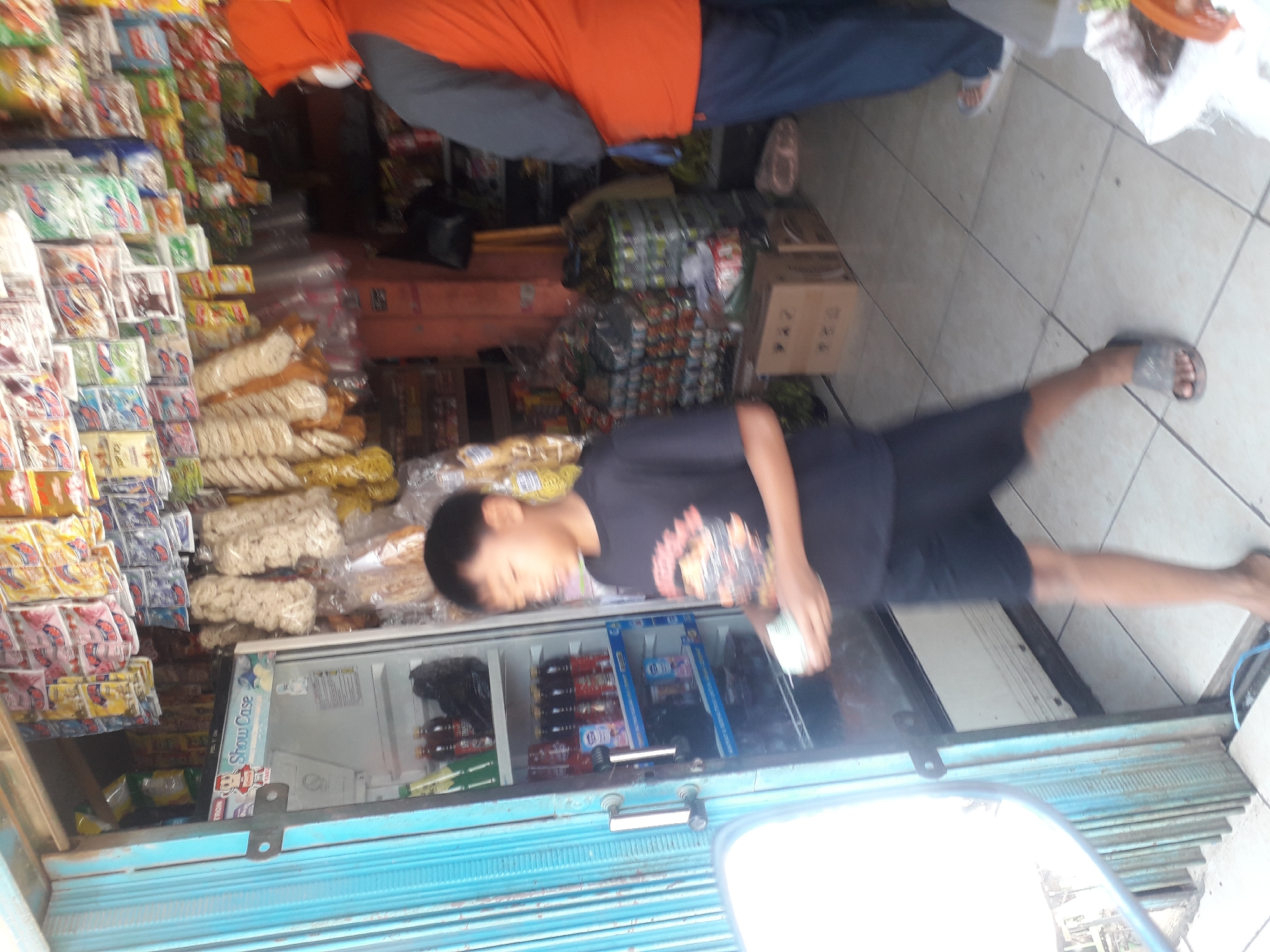 This image depicts an outdoor scene in front of a small shop in Cankuang, Indonesia. A young boy, wearing a black t-shirt and dark shorts, is walking towards the right of the frame, holding a light-colored object in his right hand. He is facing slightly away from the viewer, with his body angled to the right. To his left, a blue metal door with vertical slats is partially visible.
Behind the boy, an open refrigerator displays several bottles of drinks. Shelves above the refrigerator are stocked with various packaged goods, including brightly colored snack bags and other retail items. To the right of the refrigerator, a person wearing an orange hooded jacket over a grey shirt and blue pants is also visible, facing away from the viewer. This person appears to be a woman, indicated by the hijab-like orange hood covering her head and neck.
The shop appears to be a small, local establishment, with merchandise displayed both inside and outside. The ground is tiled with light-colored square tiles. Stacks of boxes and bags of goods are visible behind the people. The overall impression is one of a busy, informal marketplace setting.