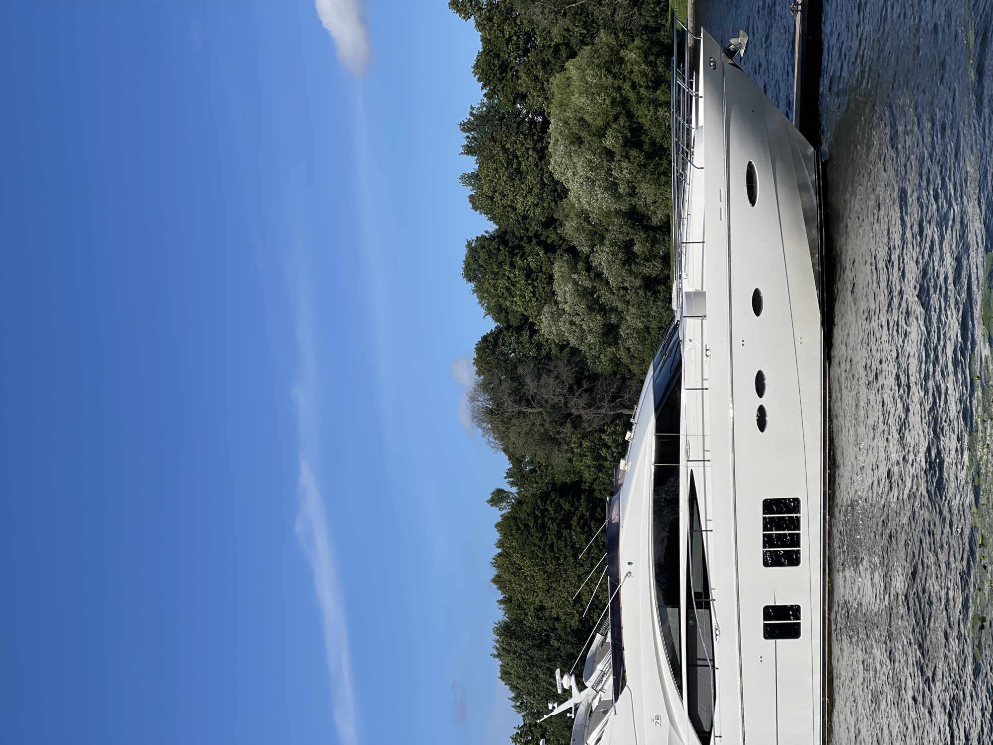 A pristine white yacht is docked on a body of water under a clear blue sky with a few wispy white clouds. The yacht, featuring sleek lines and large dark windows, is positioned parallel to a wooden dock. The background is dominated by a dense line of lush green trees, creating a natural backdrop. The water is a deep blue-green, with gentle ripples visible on its surface. There are no people visible in the image.