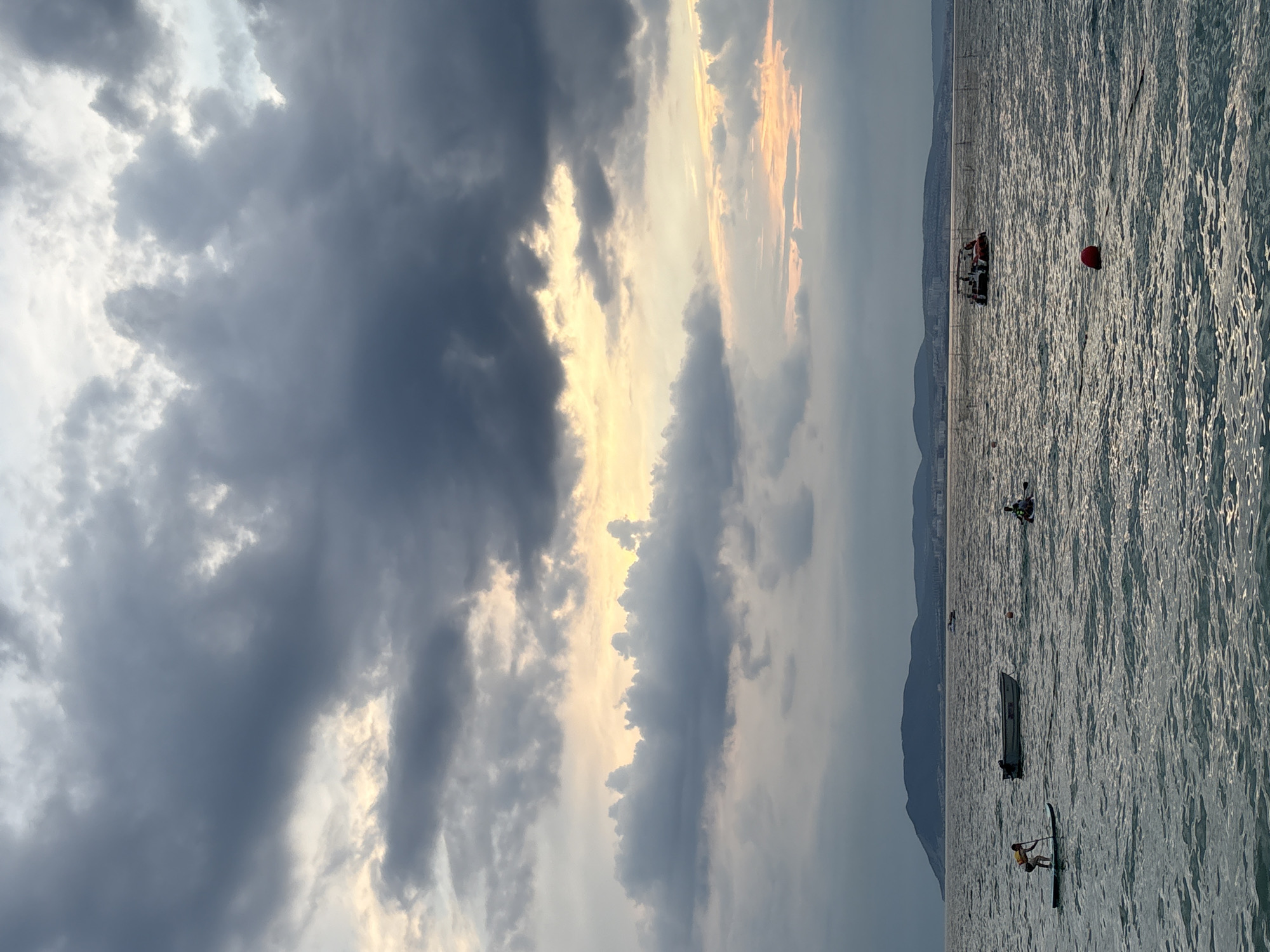 The image depicts a view of the sea in Novorossiysk, Russia, likely near the embankment or a recreational area. The sky is filled with dramatic, dark clouds, with patches of pale yellow and orange light breaking through, suggesting either sunrise or sunset. The sea is moderately choppy.
In the foreground, on the water, there are several objects and people:
* A person on a paddleboard, wearing a yellow vest, is paddling away from the viewer.
* A small, light-colored boat is anchored or drifting in the water.
* Further out, another person is in a kayak or similar small watercraft, paddling.
* A red buoy is floating in the water.
* A larger boat, possibly a pleasure craft with several people on board, is visible in the middle ground.
In the background, a mountainous coastline with a city or town nestled along it stretches across the horizon. The overall atmosphere is somewhat moody due to the cloudy sky, but the presence of water activities suggests a degree of pleasant weather. The scene appears to be a candid shot capturing a moment of activity on the water under a dynamic sky.