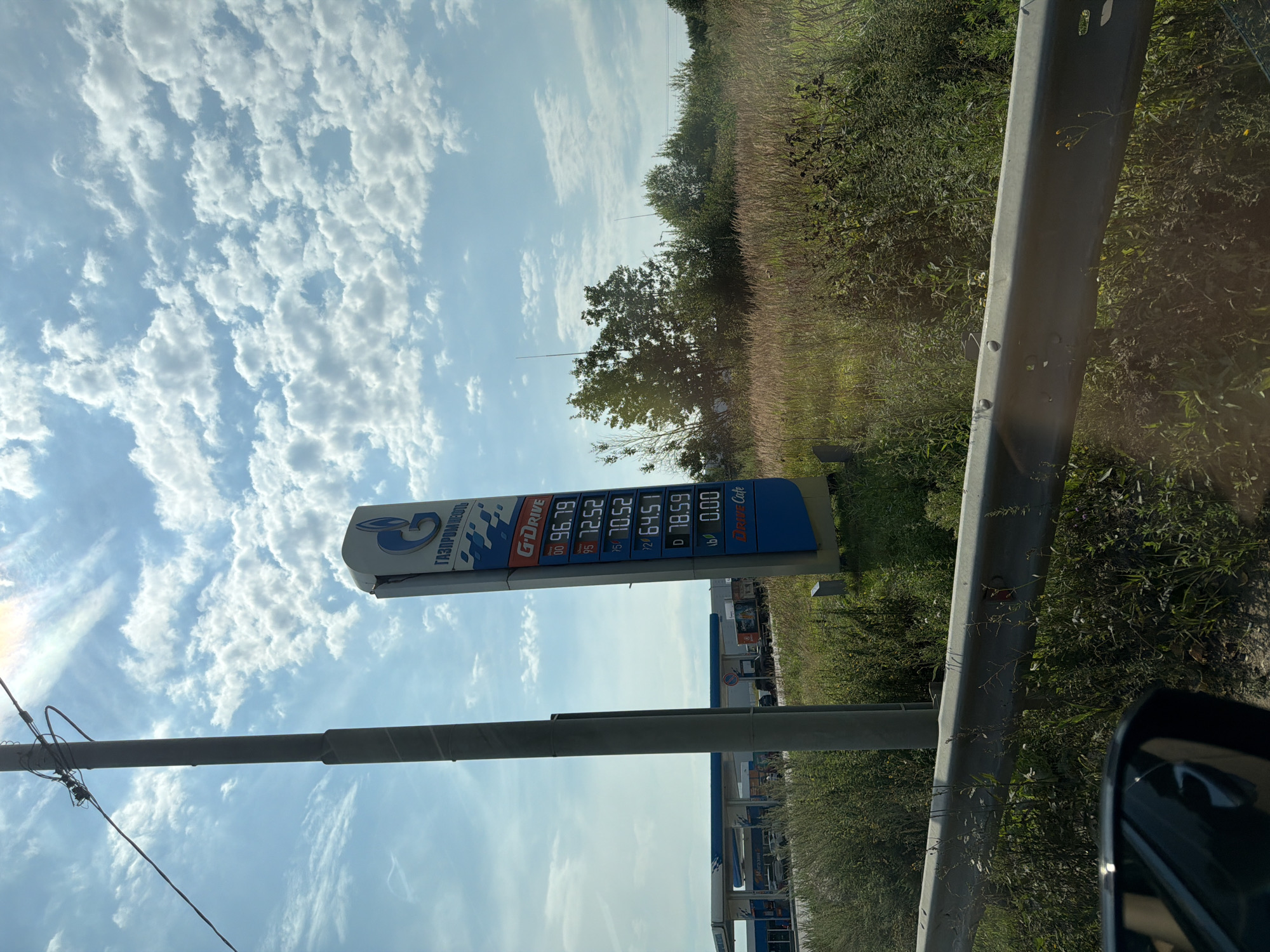 The image shows a gas station sign in Durykino, Russia. The sign displays fuel prices including G-Drive, 96.79, 95 72.52, 92 70.52, 76 64.51, D 78.59, and 166 0.00. There is also a "Drive Cafe" advertised. In the background, a gas station canopy with pumps is visible, along with a road and some trucks. The scene is set against a bright blue sky with fluffy white clouds and is surrounded by overgrown vegetation. The photograph is taken from a car, with a car's side mirror visible in the lower left corner.