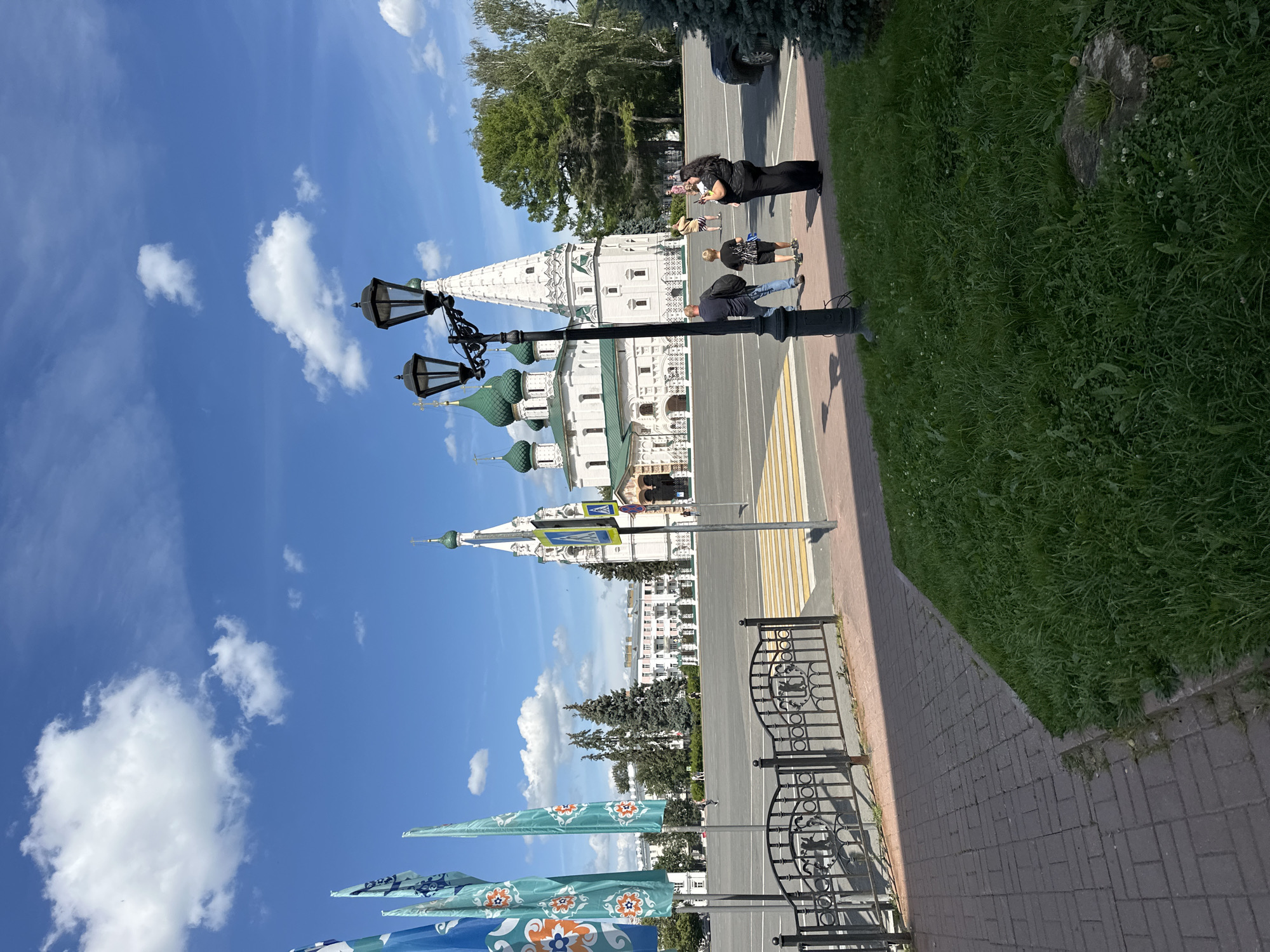 This is a ground-level, eye-level, outdoor, daytime, vertical, full-shot photograph taken on a clear, sunny day with scattered clouds in the sky. The scene captures a public square with a prominent white church complex featuring green onion domes and a tall, white bell tower with intricate architectural details in the background. In the mid-ground, a few individuals are walking across a crosswalk towards the church. To the left of the church are several tall, slender flags with a blue and orange floral pattern. In the foreground, there is a paved walkway with a brick pattern, a patch of green grass with a single stone, a black metal fence, and a black lamppost with two lights. The overall impression is of a serene and historical urban environment.