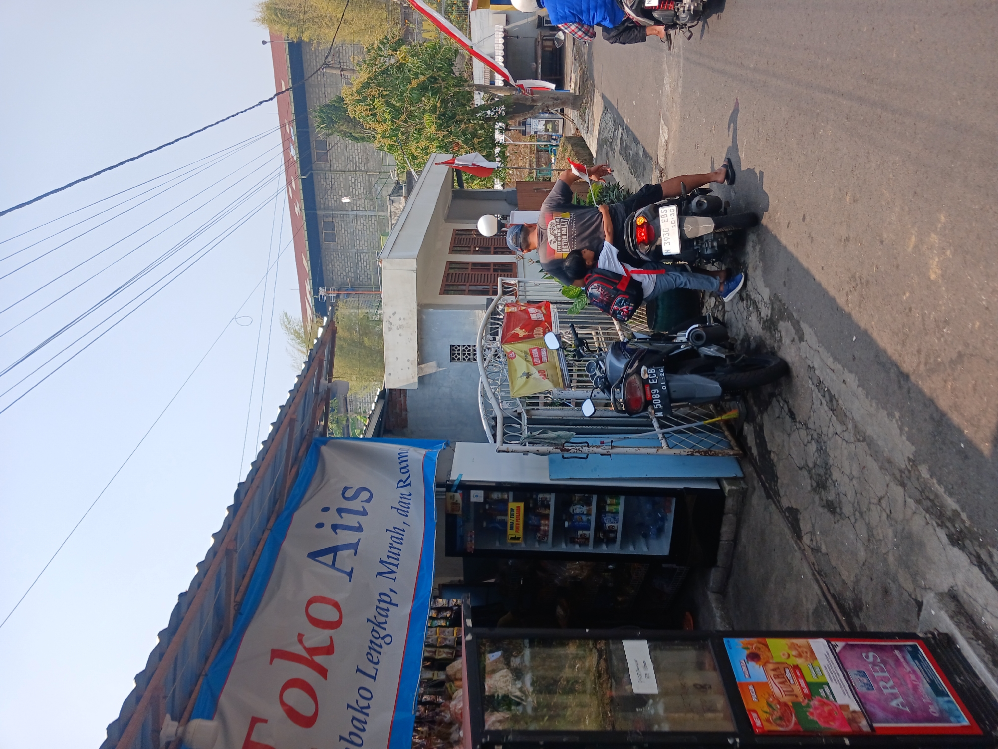 This is a street-level, eye-level, medium shot photograph taken in Watugede, Indonesia. The image captures a street scene in front of a small shop. A banner above the shop entrance reads "Toko Aiis" and below it, in smaller text, "bako Lengkap, Murah, dan Rame". The shop appears to be a convenience store with a glass-fronted refrigerator displaying various drinks. To the left of the shop entrance, another glass-fronted display case is visible, filled with goods. A person is standing in the doorway of the shop.
In the foreground, two motorcycles are parked. On the left motorcycle, a person's legs are visible. On the right motorcycle, a man is seated, wearing a t-shirt with the text "INDONESIA RETRO RALLY" and a blue cap. A child wearing a backpack is standing behind him, with their arms around his waist. The man is holding a small Indonesian flag. Another motorcycle is visible further down the street, with a person wearing a blue vest on it.
The environment appears to be a rural or semi-urban area with a paved road. Buildings are visible in the background, including a multi-story structure with a red roof. Trees and bamboo are also present. Several Indonesian flags are displayed on poles and strung across the street, suggesting a national celebration or event. The sky is clear and bright. The overall impression is a snapshot of everyday life in a small Indonesian town.