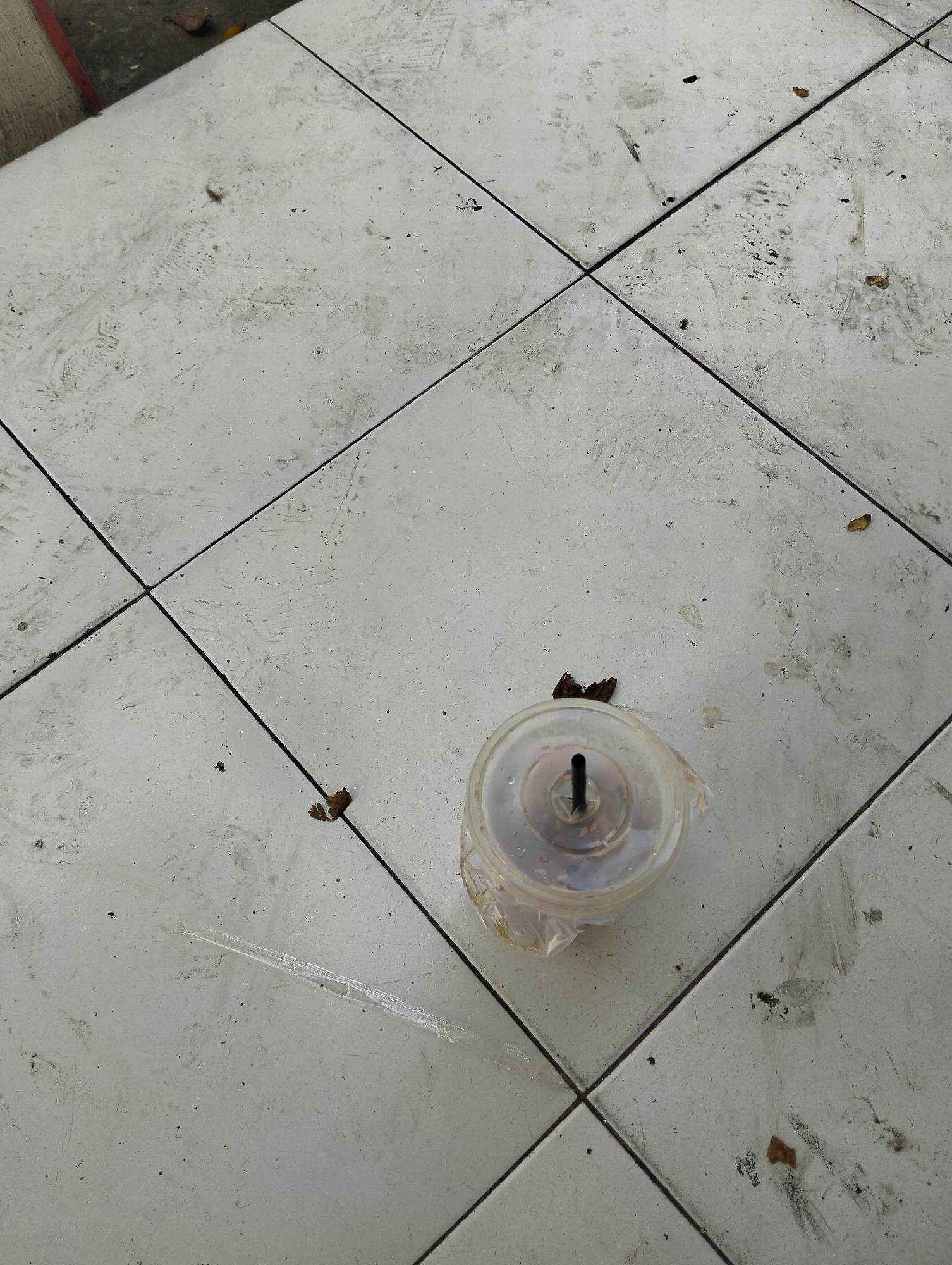 The image shows a close-up, high-angle view of a tiled floor in an outdoor setting in Surabaya, Indonesia. The tiles are light-colored and laid in a grid pattern, with visible grout lines. The floor is dirty, with numerous scuff marks, footprints, and scattered debris including dry leaves and small pieces of trash. In the lower center of the image, a clear plastic cup with a black straw is lying on its side, partially covered by a plastic wrapper. The cup appears to contain some liquid and possibly remnants of a drink. To the left of the cup, a single plastic straw is also visible on the ground. The scene suggests a public area that has experienced recent foot traffic and a lack of cleaning.