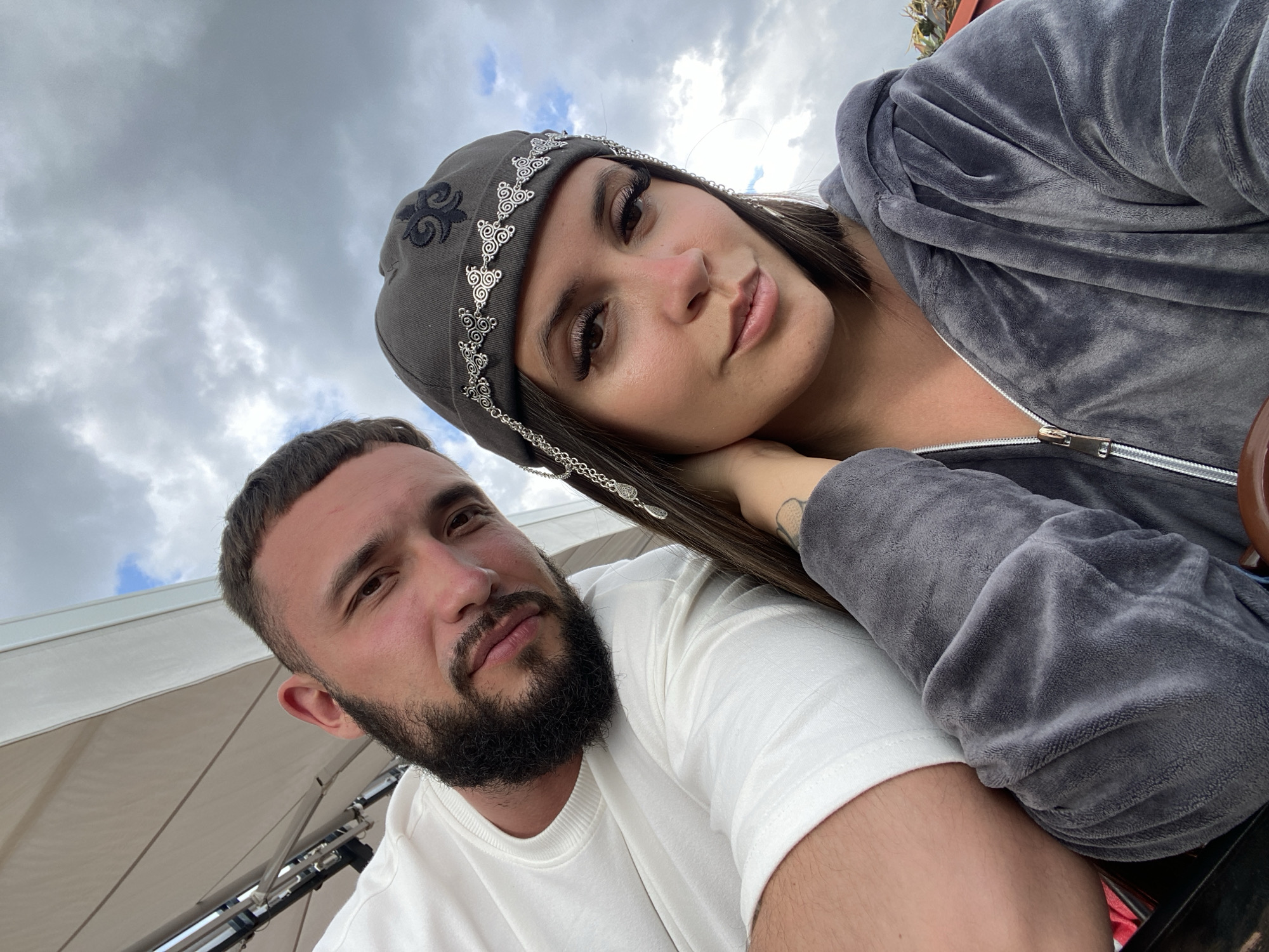A close-up, selfie-style photo shows a man and a woman outdoors. The man, on the left, is wearing a white t-shirt and has short dark hair and a beard. The woman, on the right, is wearing a gray hooded jacket and a gray beanie with silver embellishments. She has long brown hair and is wearing makeup. They are both looking at the camera. The background consists of a cloudy sky and what appears to be a white awning or canopy structure. The photo appears to have been taken in Gunib, Russia, a mountainous village known for its picturesque scenery.