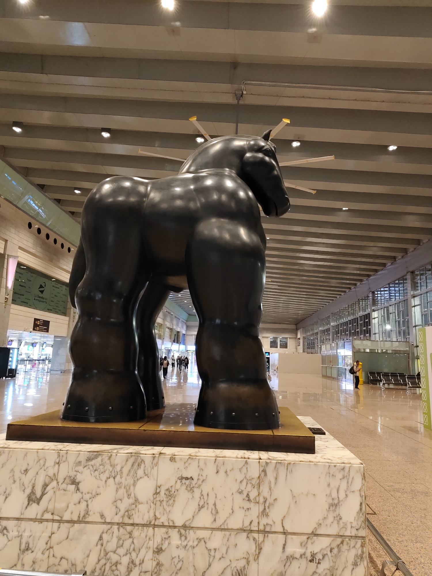 A large, dark, rotund horse sculpture, characteristic of Fernando Botero's style, stands prominently on a white and grey marble-patterned base with a golden-hued top. The sculpture is viewed from its rear-right, showcasing its exaggerated, voluminous form.
The scene is set in an expansive, well-lit indoor public space, resembling an airport terminal or large transportation hub in El Prat de Llobregat, Spain. The ceiling features exposed concrete beams and numerous recessed lights, some casting bright glares.
In the background, several people are walking through the concourse. On the left, a light blue-green panel displays Spanish text, including "Las Tiendas del Aeropuerto" (The Airport Shops) with a right-pointing arrow, and another directional sign below it. Further to the right, a lone person stands near rows of empty black waiting benches adjacent to large windows. A repeating decorative pattern of "8888" is visible on a pillar or wall on the far right. The floor is light-colored and reflective, suggesting an open and modern interior. The setting appears to be during daytime.