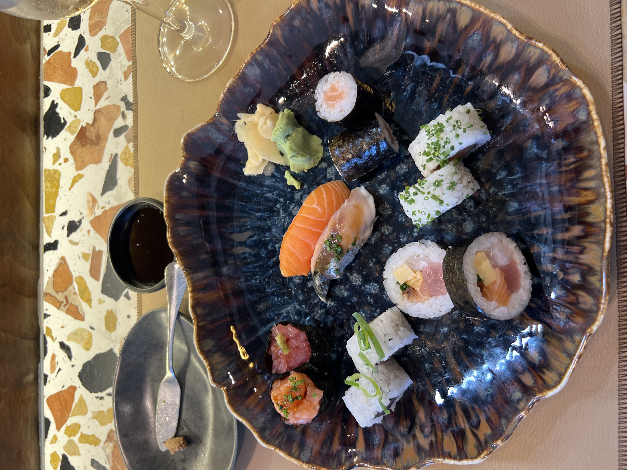 A high-angle, eye-level shot captures a plate of sushi in a restaurant in Porto, Portugal. The sushi is arranged on a dark blue, organically shaped plate with brown and gold accents, sitting on a tan placemat. The plate contains various types of sushi including nigiri (salmon and other fish varieties), maki rolls with chopped chives, and rolls with scallions. Accompanying the sushi is a small mound of wasabi and pickled ginger. To the left of the plate, there is a small, dark-colored dipping sauce container, a silver spoon or small serving utensil resting on a smaller, gray-colored plate. Part of a wine glass is visible in the upper left corner. The table also features a terrazzo countertop with a mix of white, gray, brown, and yellow fragments. The setting suggests an upscale or modern dining environment.