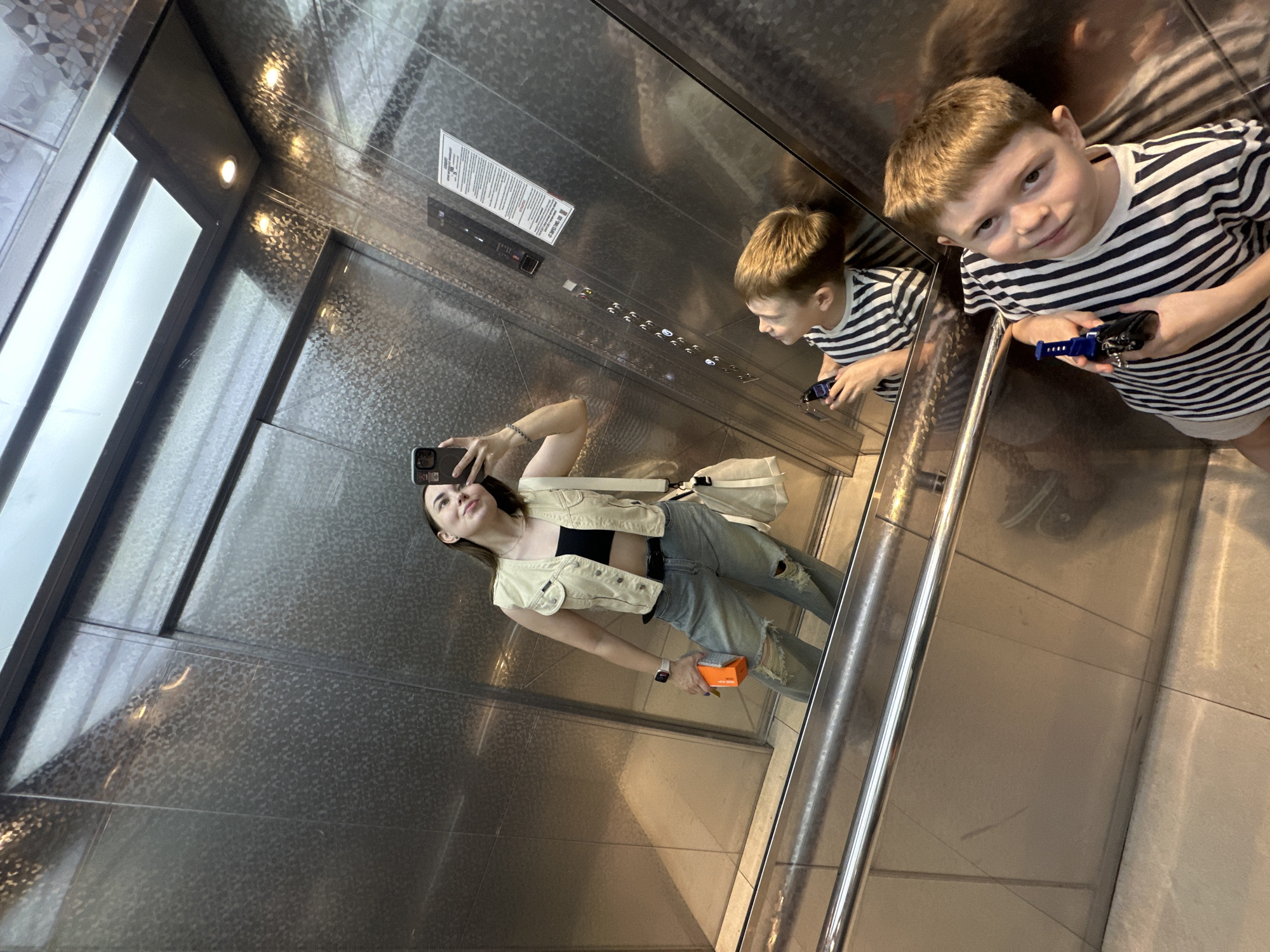 An indoor, high-angle shot shows a woman and a boy in an elevator. The elevator has a mirrored finish, reflecting the subjects and the interior. The woman holds a smartphone, presumably taking the picture. She wears a black top, jeans with holes, and a light-colored vest with a white purse hanging off of her shoulder. She is also holding an orange box. The boy, standing on the right, wears a striped shirt and holds a small blue and black toy. A silver rail runs vertically on the right side of the frame. The scene is located in Perm, Russia, evidenced by text on the orange box.