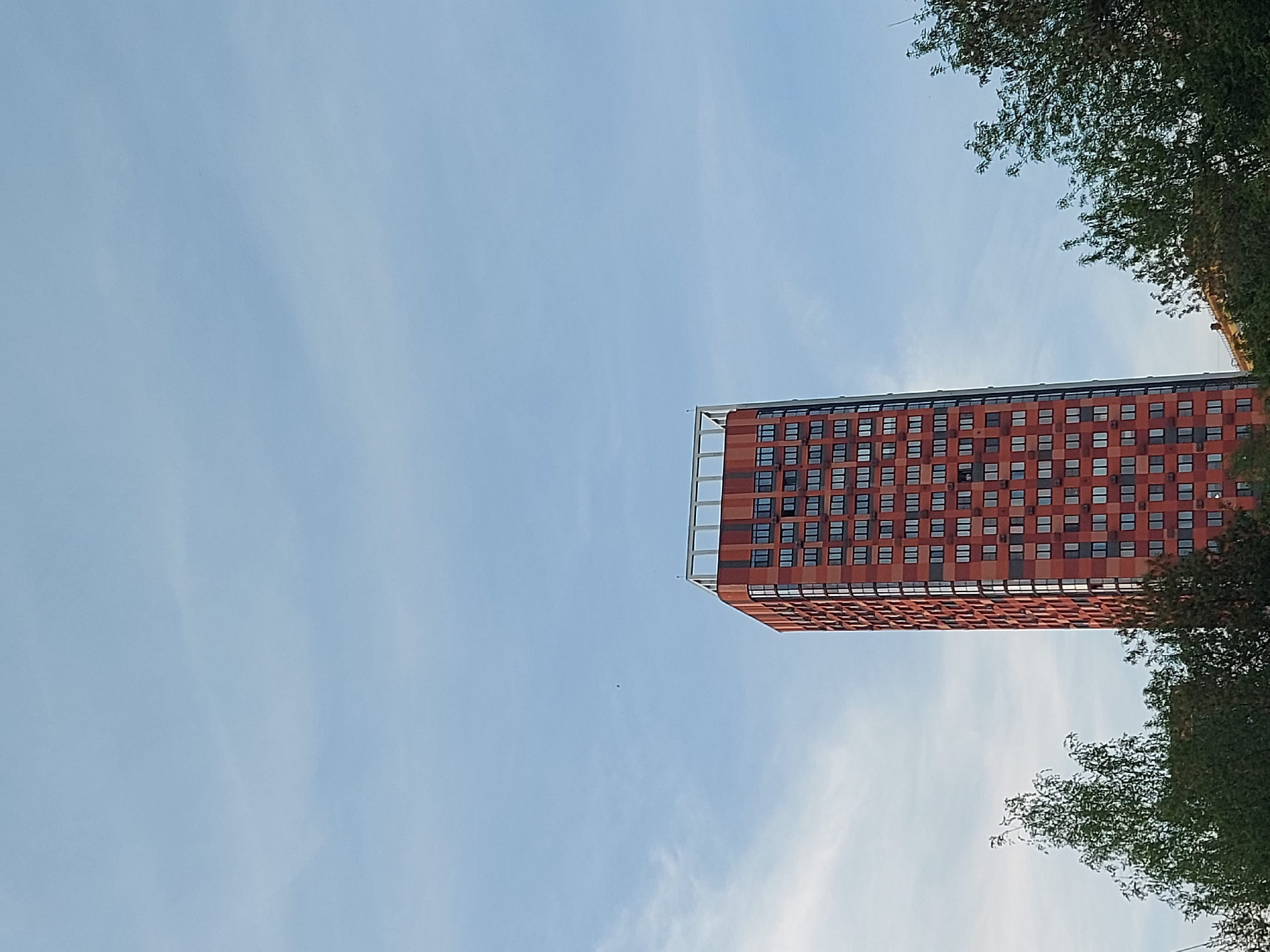 Here's a description of the image:
The image depicts a tall, red-brick building against a backdrop of a light blue sky with scattered clouds. The building has a rectangular shape with many windows evenly spaced across its facade. A white structure is visible on top of the building. Trees are partially visible on the right side of the image, framing the building and the sky. The angle of the photo suggests the building is being viewed from below, looking upwards. Based on location data, the building is likely located in the city of Izhevsk, Russia.