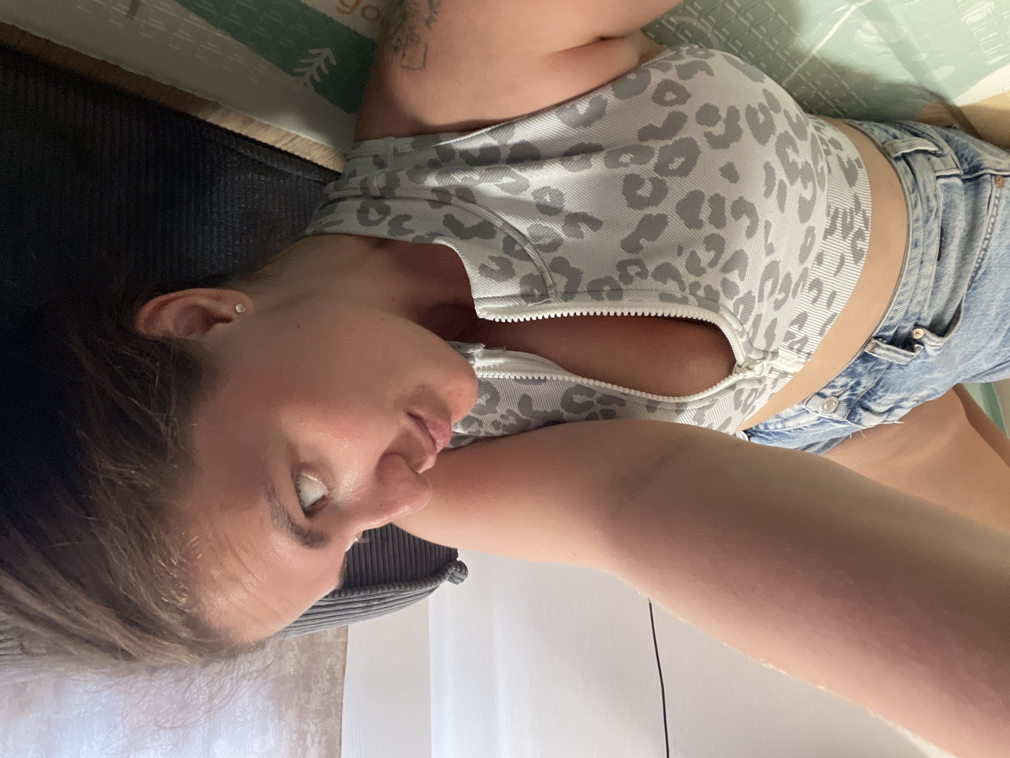 A woman is lying on her back. She is wearing a light blue and gray leopard print crop top and light blue denim shorts. She has an earring in her left ear. The background includes a black mat and a light green and white patterned surface. It is not possible to determine the location of Misaylovo, Russia from the image.